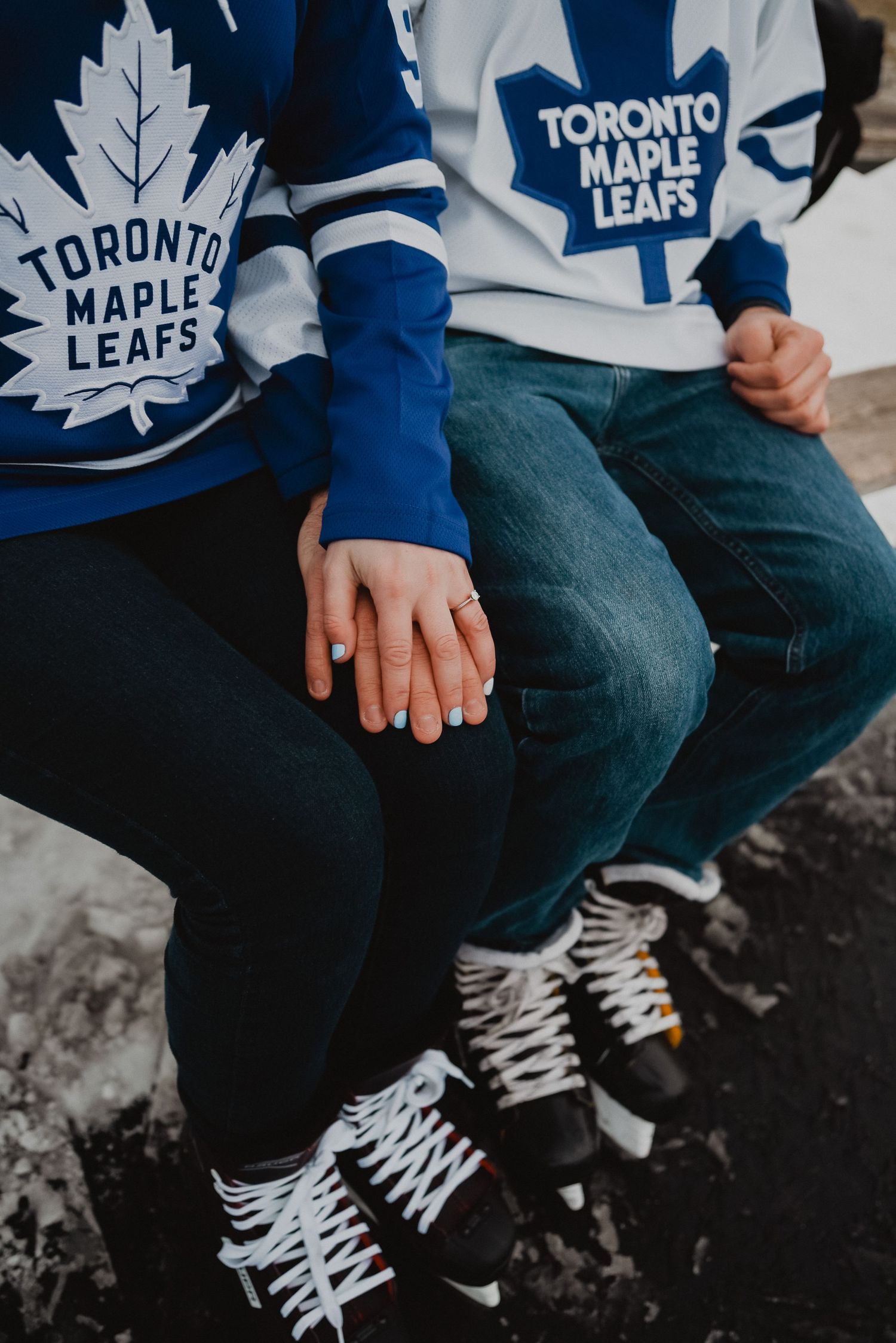 Couple wearing matching Toronto Maple Leafs jerseys holding hands while sitting in ice hockey skates.