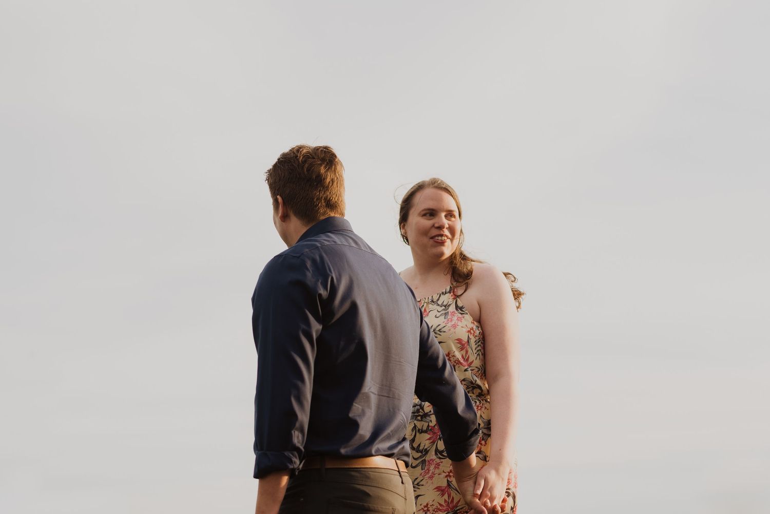 Couple sharing a tender moment outdoors, woman in floral dress smiling at man in navy shirt against overcast sky.