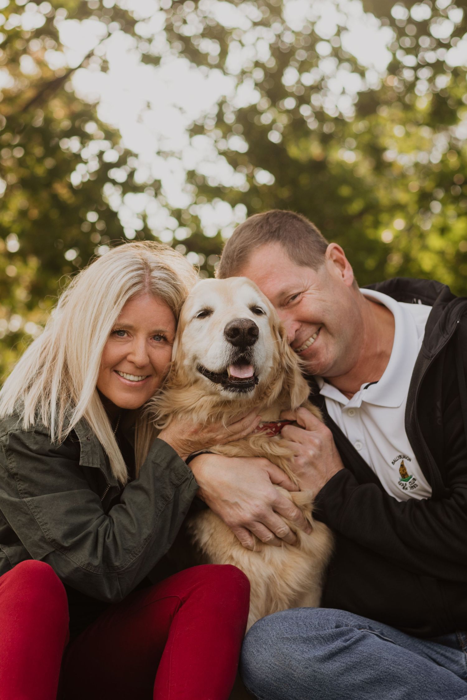 A smiling couple poses outdoors with a happy golden retriever dog, surrounded by lush green bokeh background.