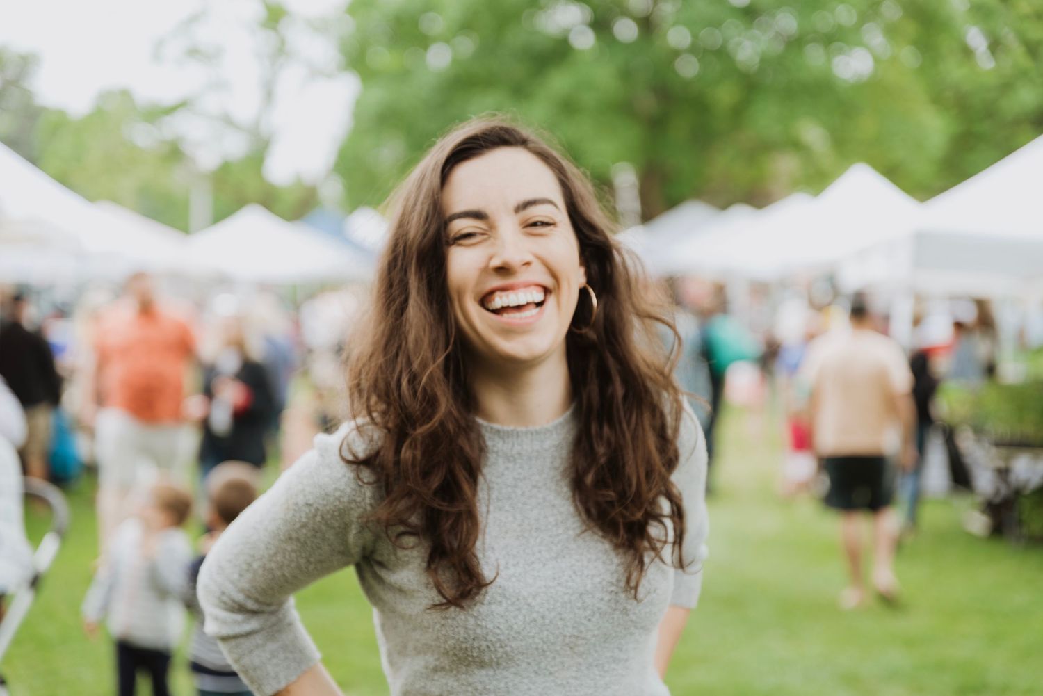 Young woman with long brown hair laughing joyfully outdoors at a summer festival market event.