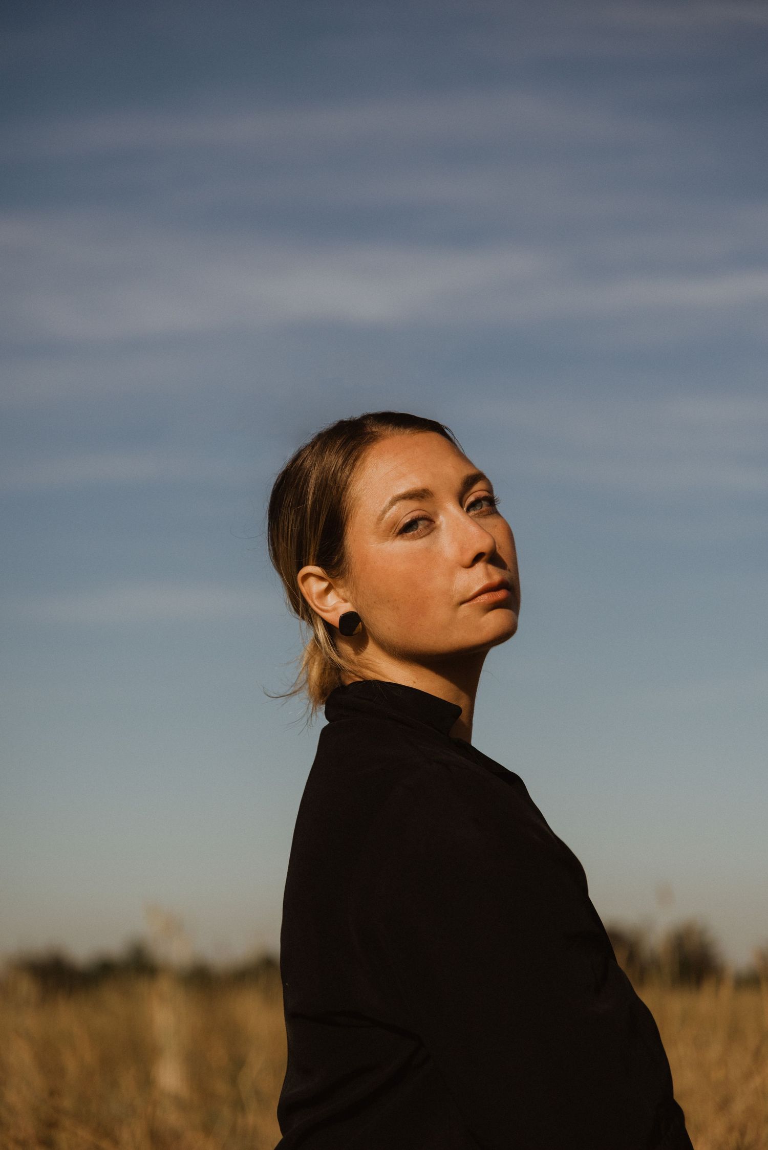 Woman in black turtleneck standing in golden field under blue sky, looking over shoulder with serene expression.