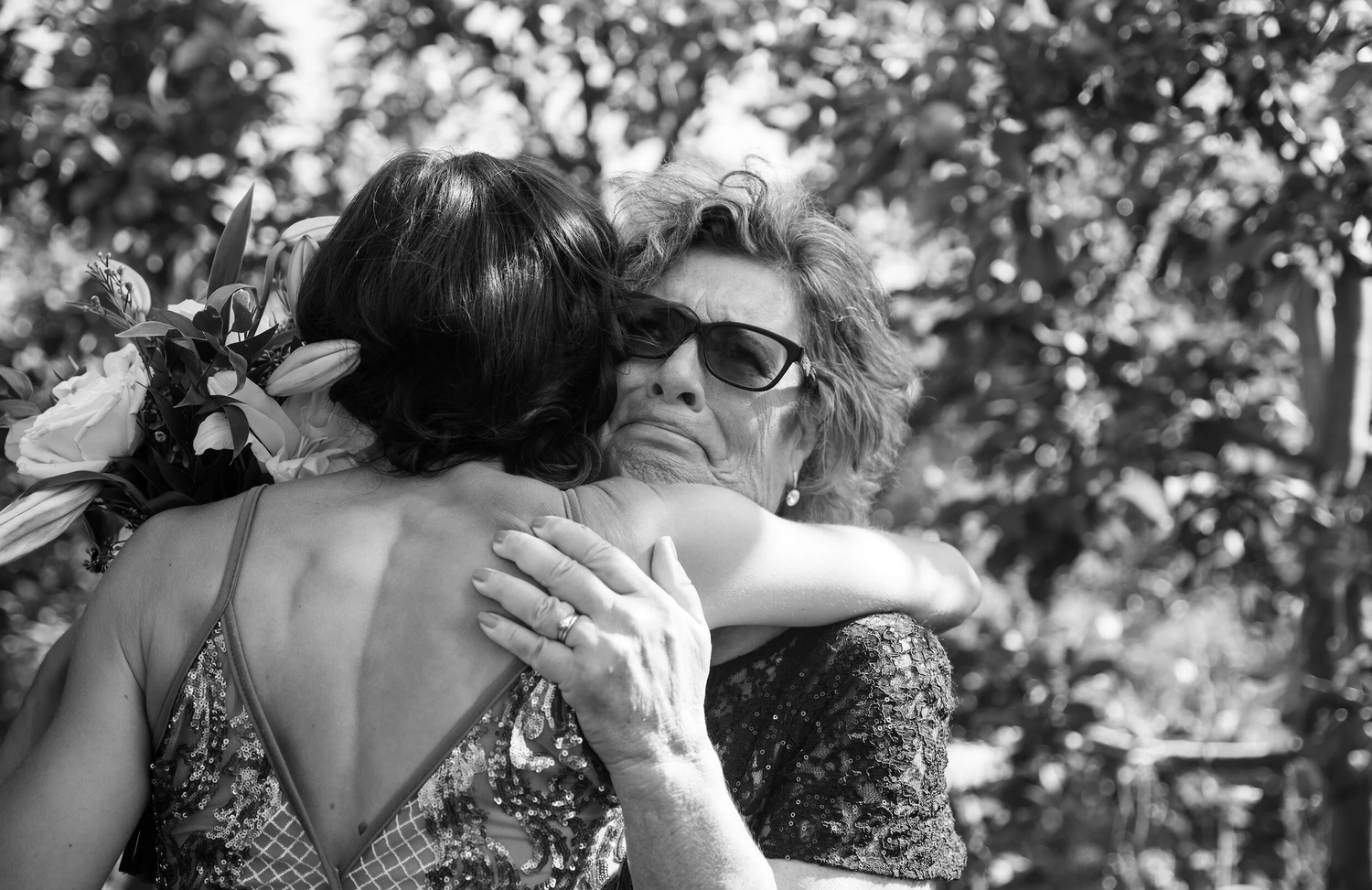 Two women share an emotional hug at an outdoor wedding, one wearing a floral accessory, in black and white.