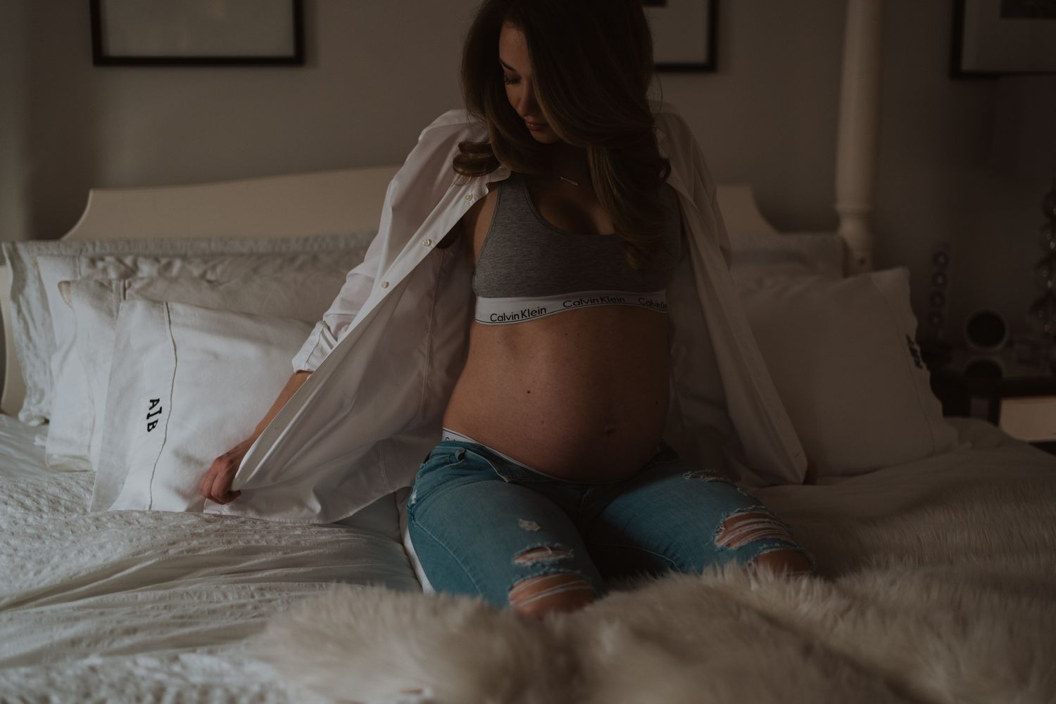 Pregnant woman in ripped jeans and gray sports bra sitting on bed, gently holding her baby bump.