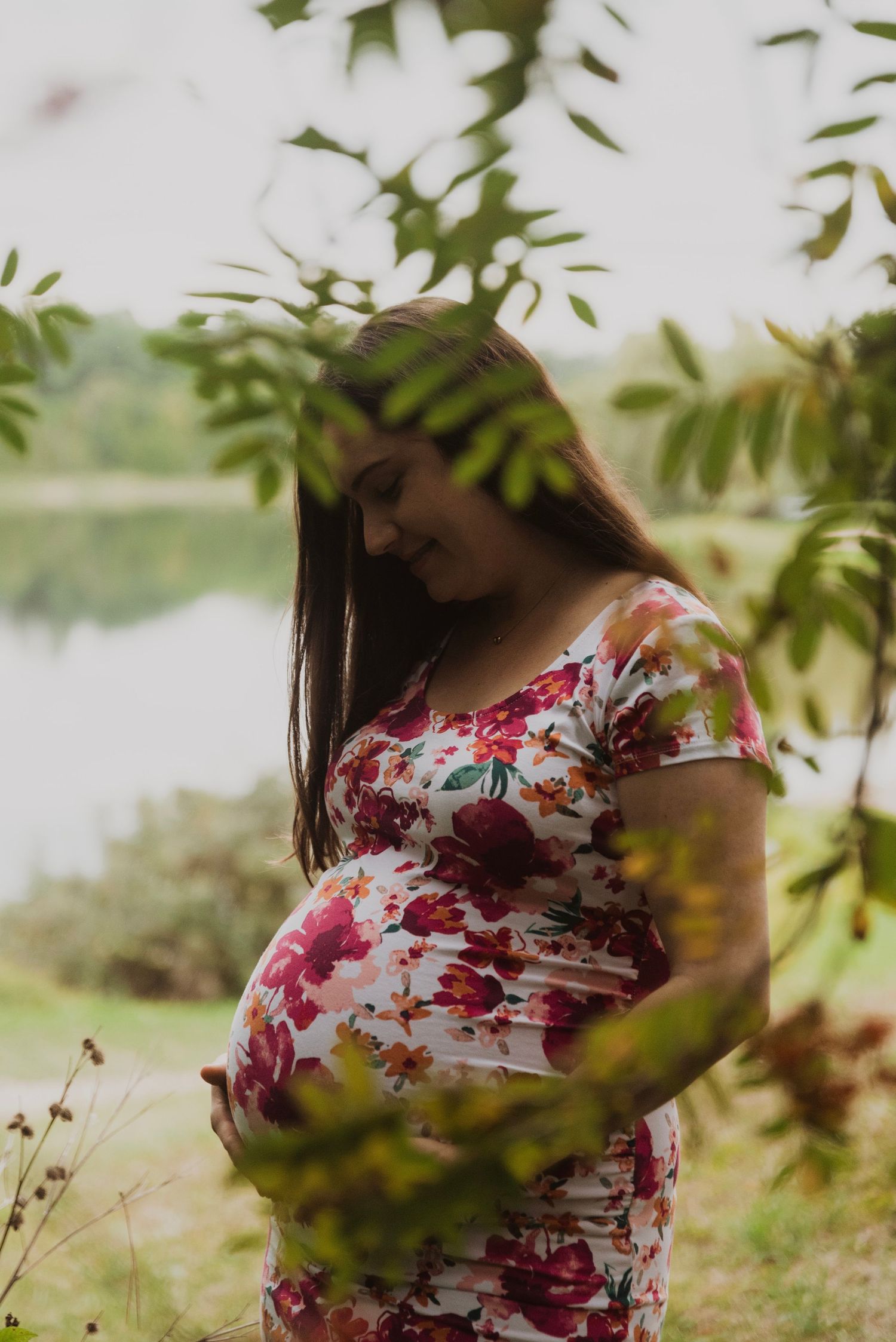 Pregnant woman in floral dress cradling baby bump outdoors near a serene lake surrounded by green leaves.