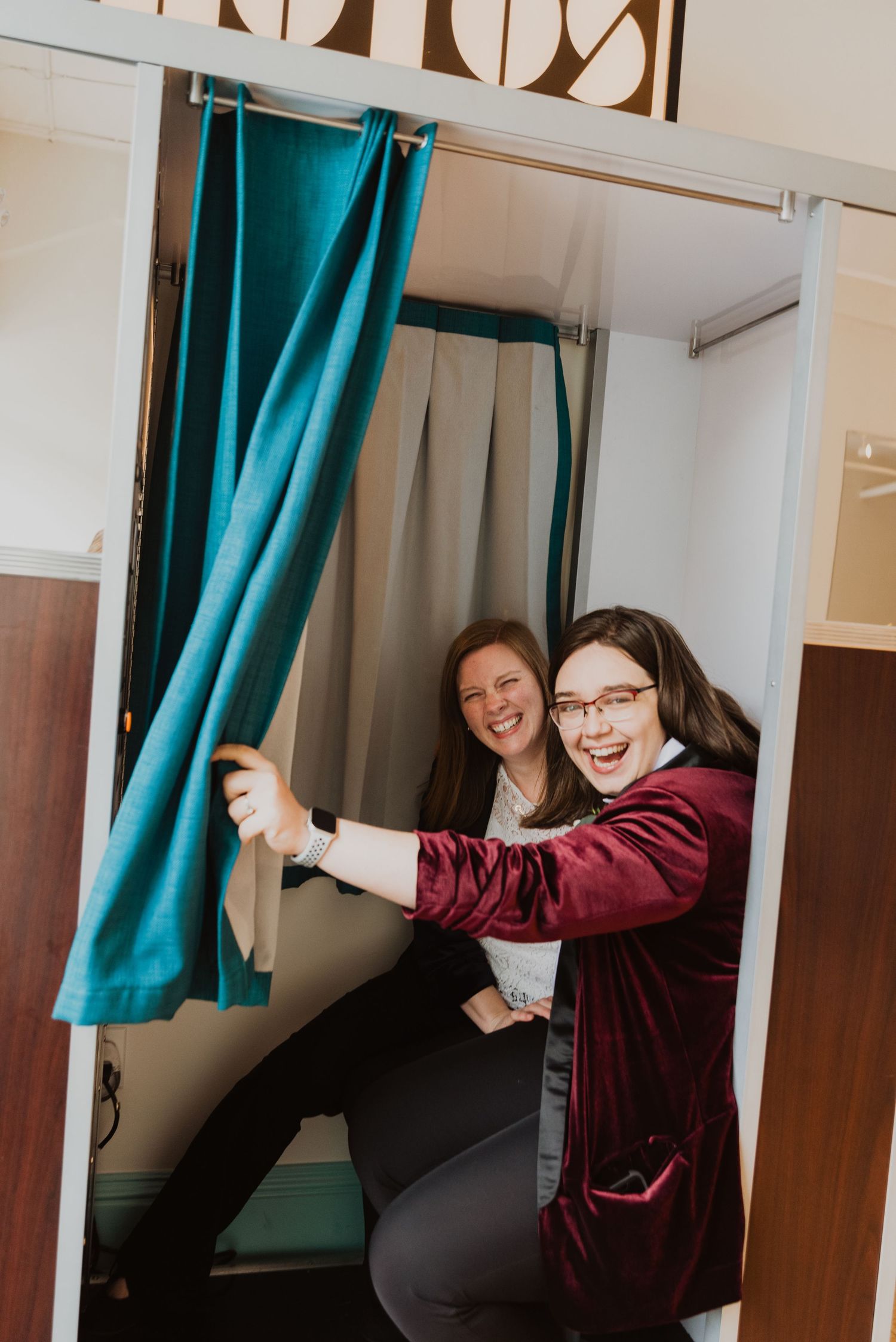 Two women laughing and posing together inside a photo booth with teal curtains.