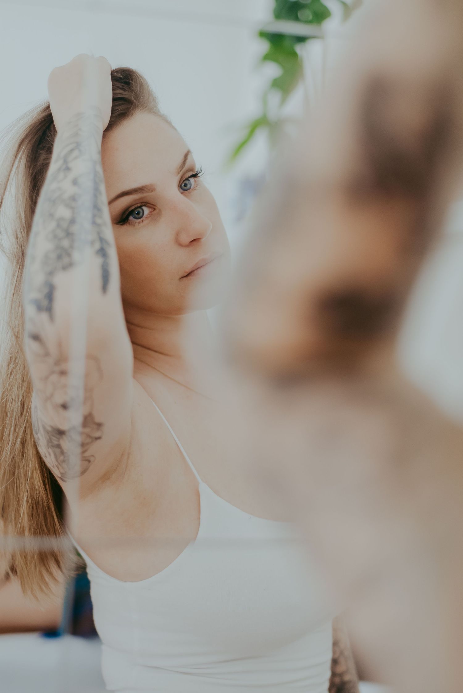 Young woman with tattoos and blue eyes in white tank top holds hair up, reflected in mirror.