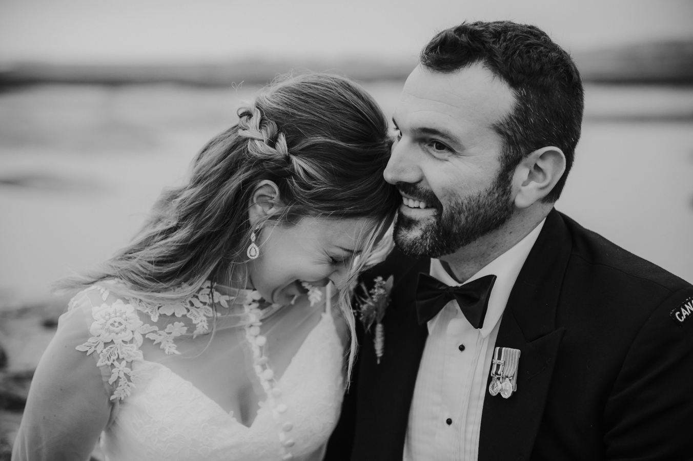 Black and white photo of a smiling bride and groom sharing a joyful moment on their wedding day near a waterfront.