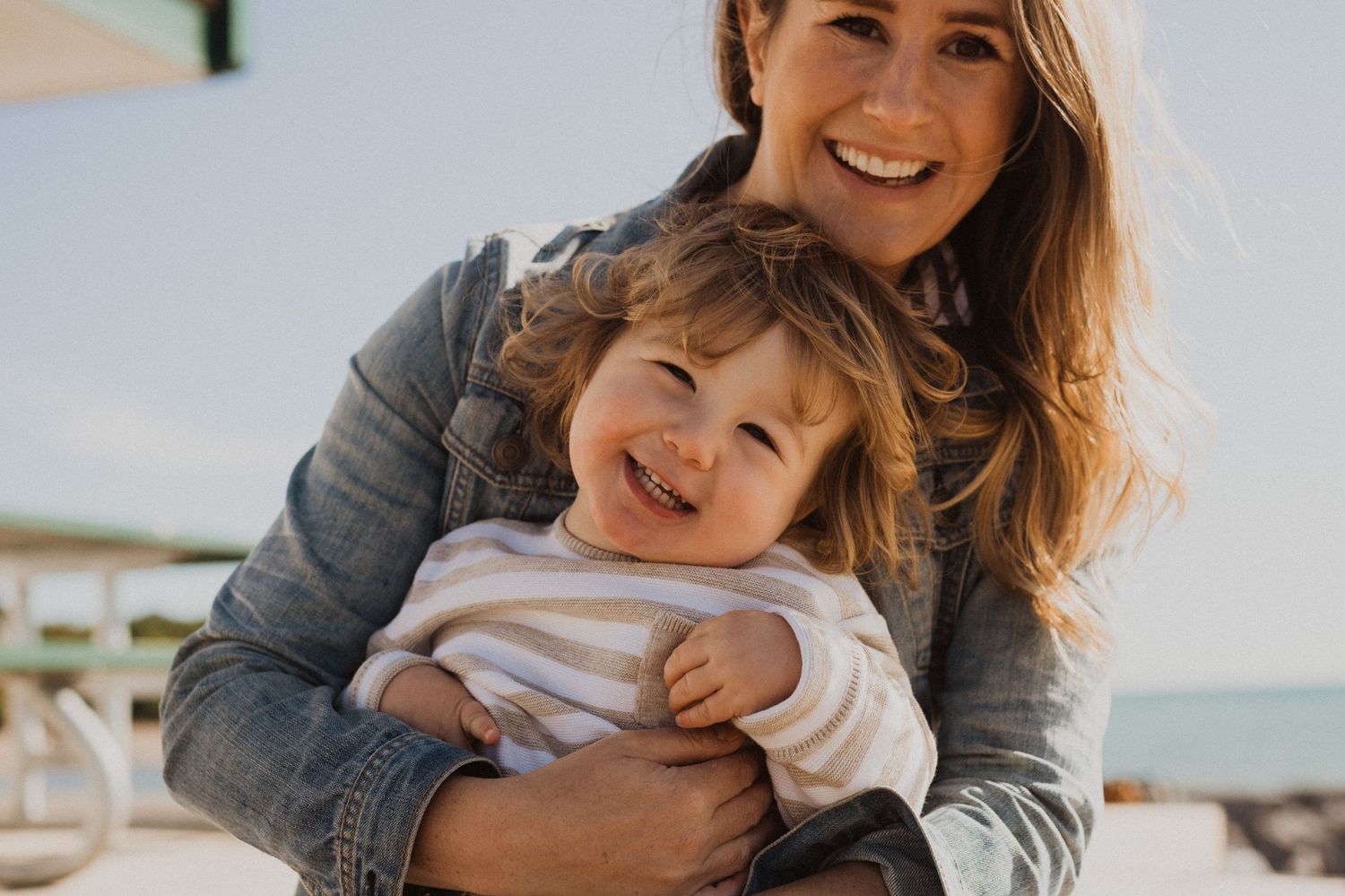 Smiling mother holding happy toddler at the beach, both wearing casual denim and white outfits on a sunny day.