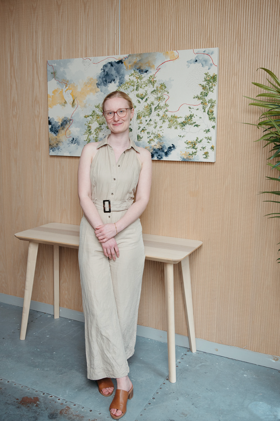 Woman in beige jumpsuit standing confidently in a modern interior with wooden furniture and abstract wall art.