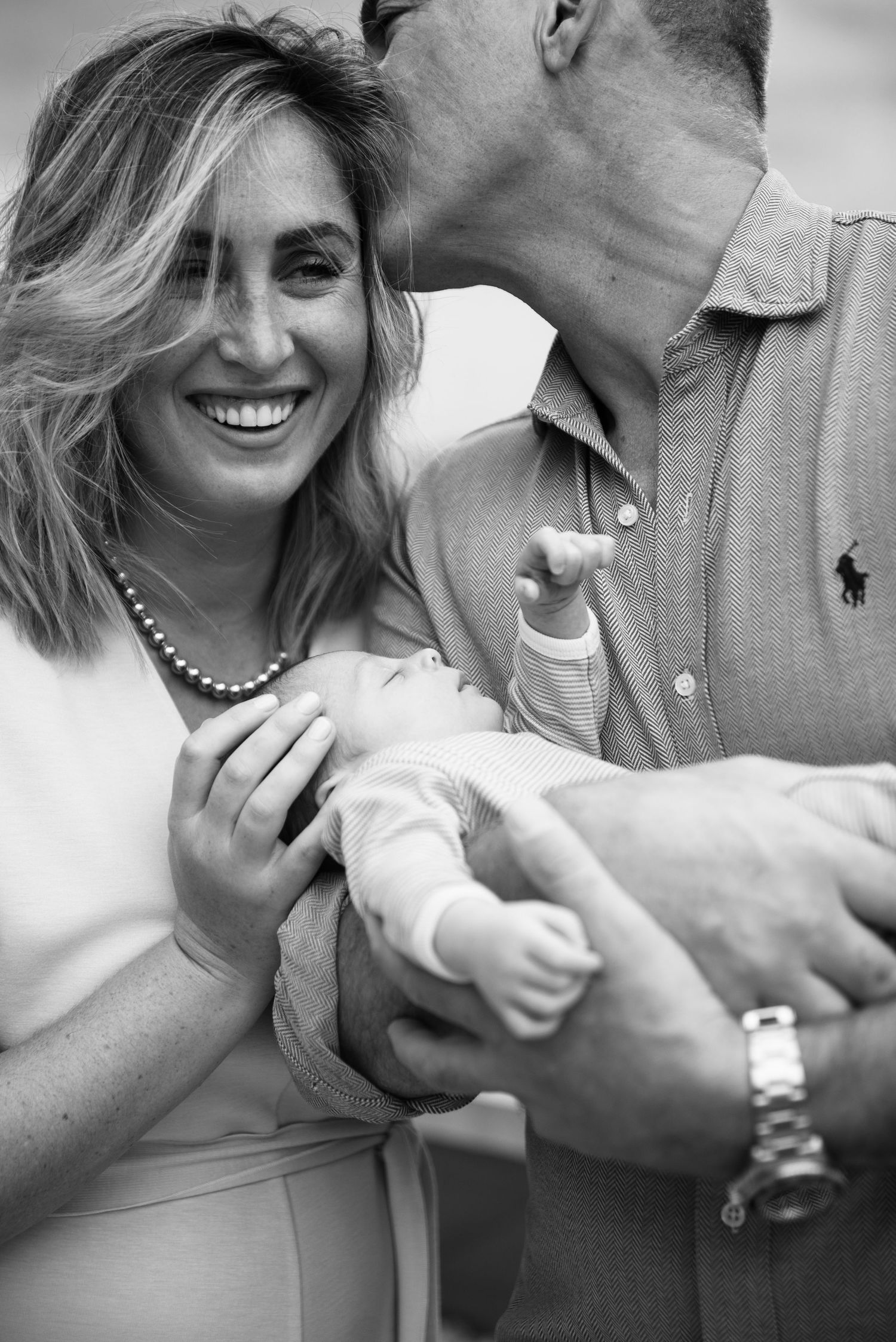 Black and white photo of smiling mother holding newborn baby while father kisses her head, family portrait.
