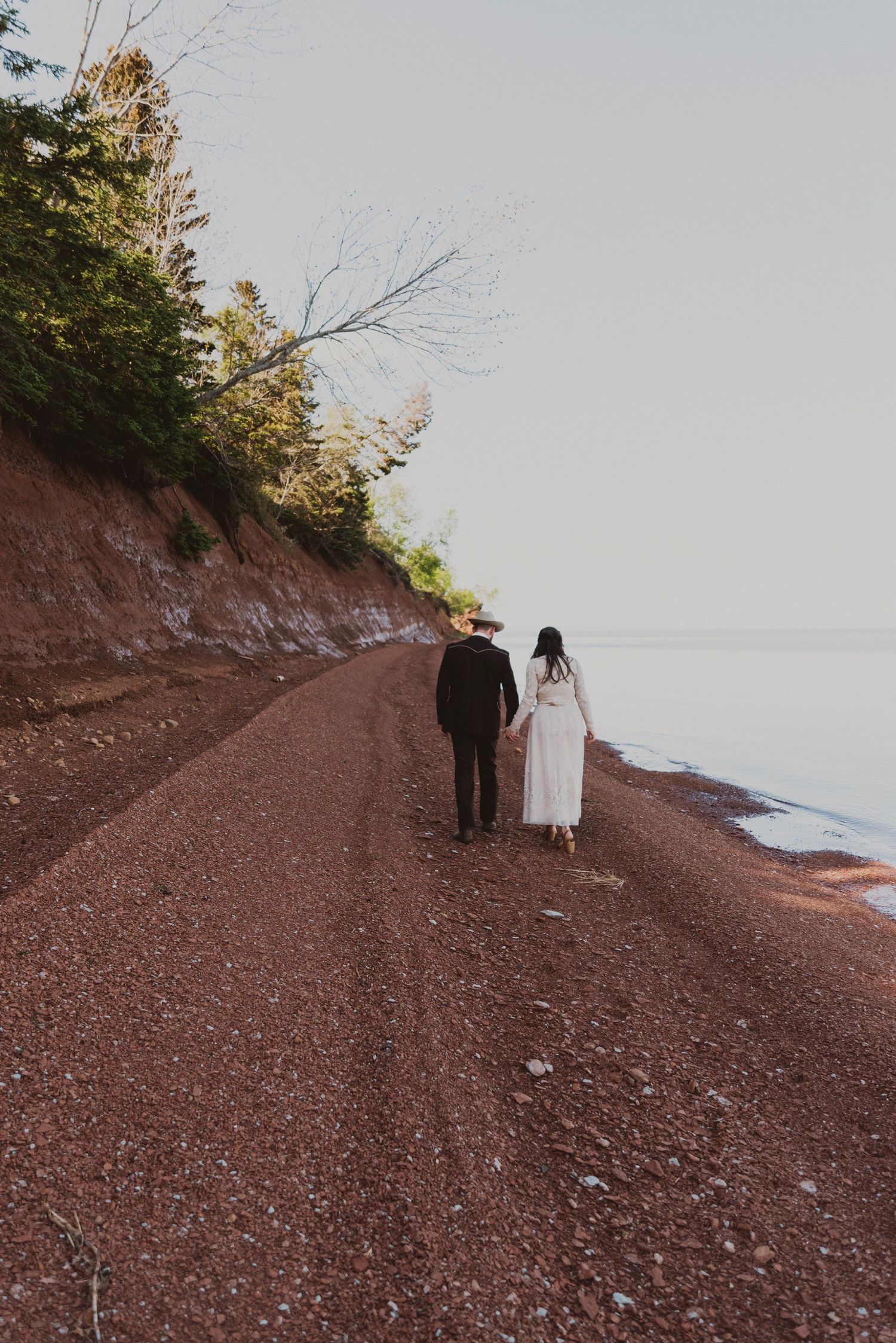 A couple in wedding attire walks hand-in-hand along a red sandy shoreline path with trees and calm water nearby.