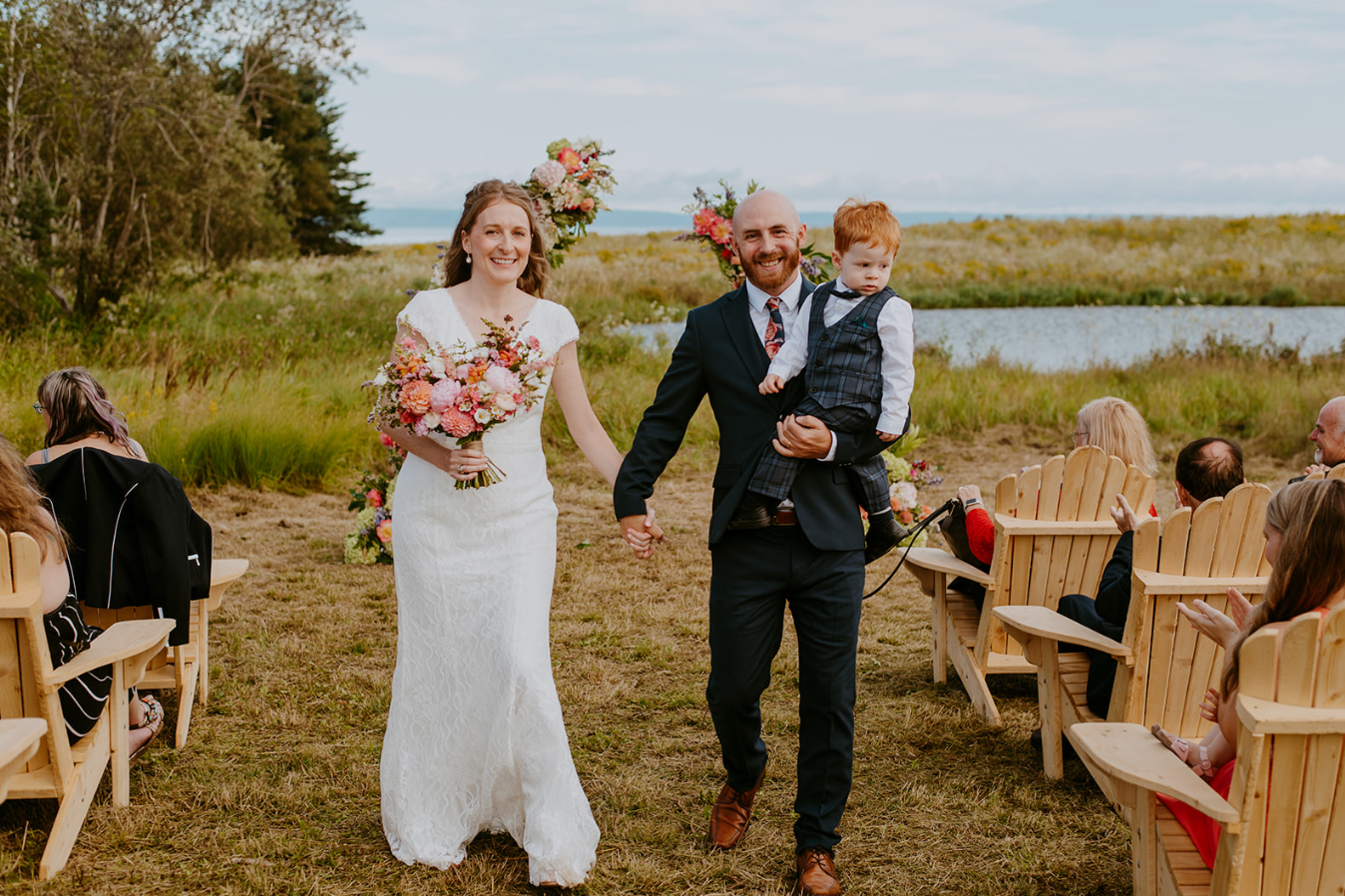 Bride and groom walk down outdoor wedding aisle holding hands, groom carries young child, waterfront backdrop.