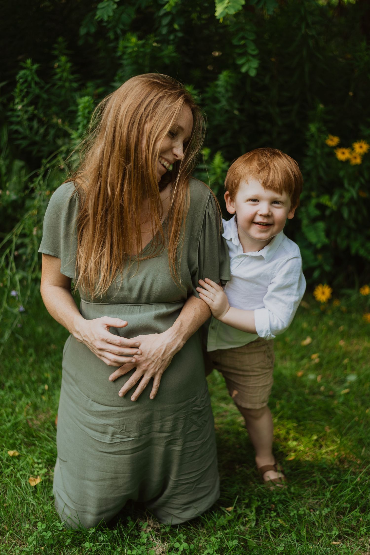 Pregnant woman in green dress kneeling outdoors with smiling red-haired toddler hugging her baby bump.