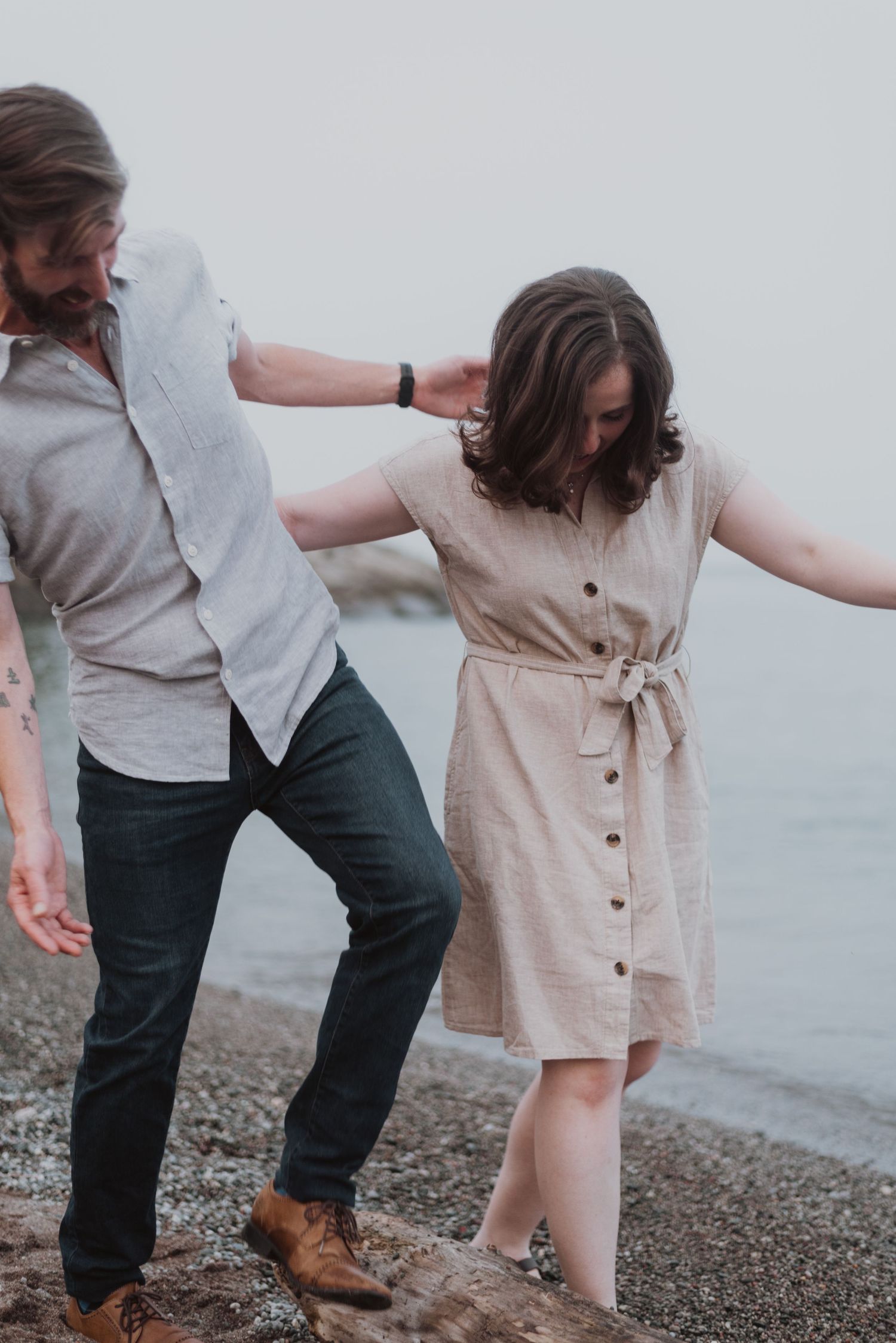 Couple balancing on rocks at beach, man steadying woman in beige dress, casual romantic moment by the water.