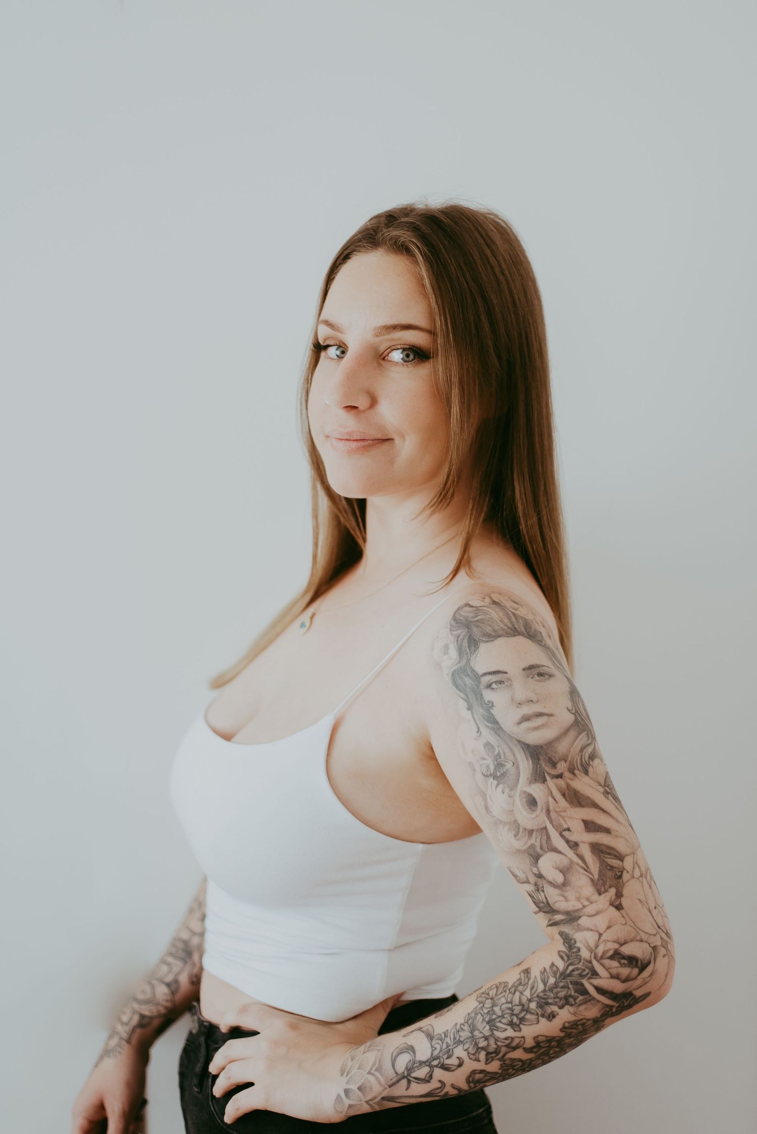 Young woman with long brown hair and full sleeve tattoo posing in white crop top against light background.
