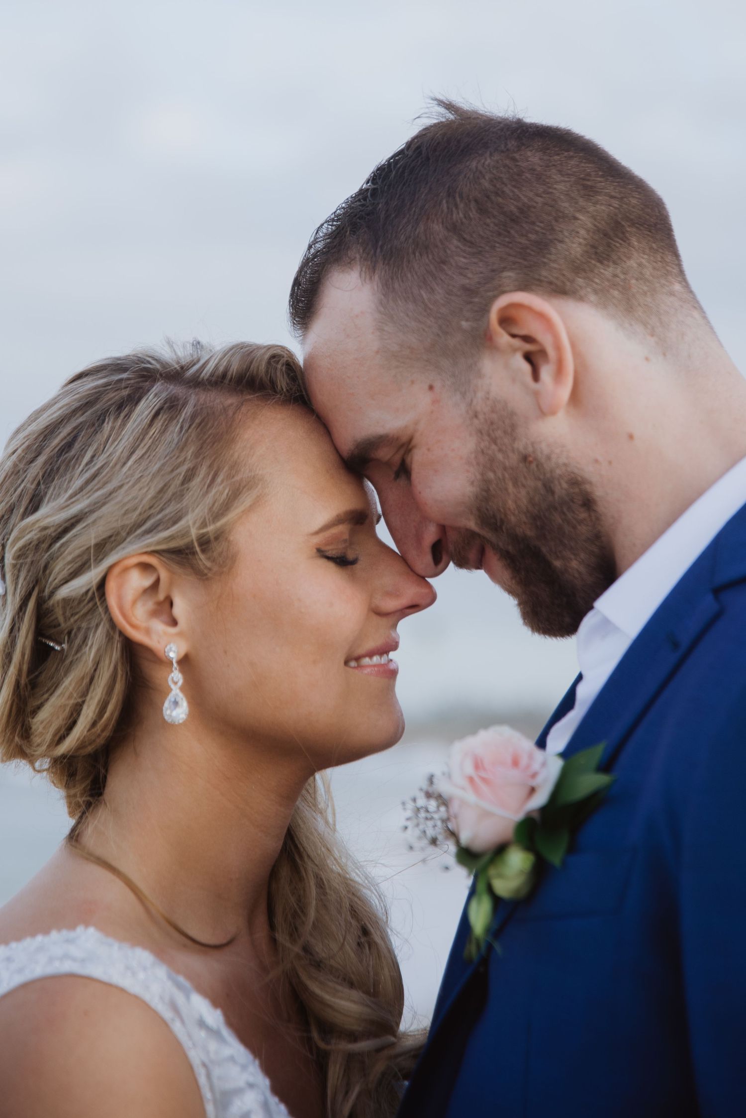 A bride and groom share an intimate forehead touch moment, with the groom in a navy suit and pink boutonniere.