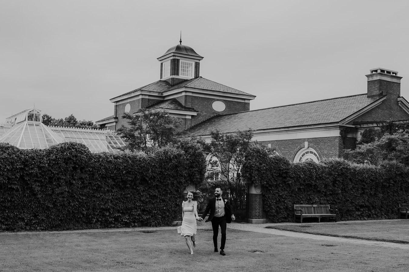 Black and white photo of a couple holding hands walking in front of a brick building with hedges.
