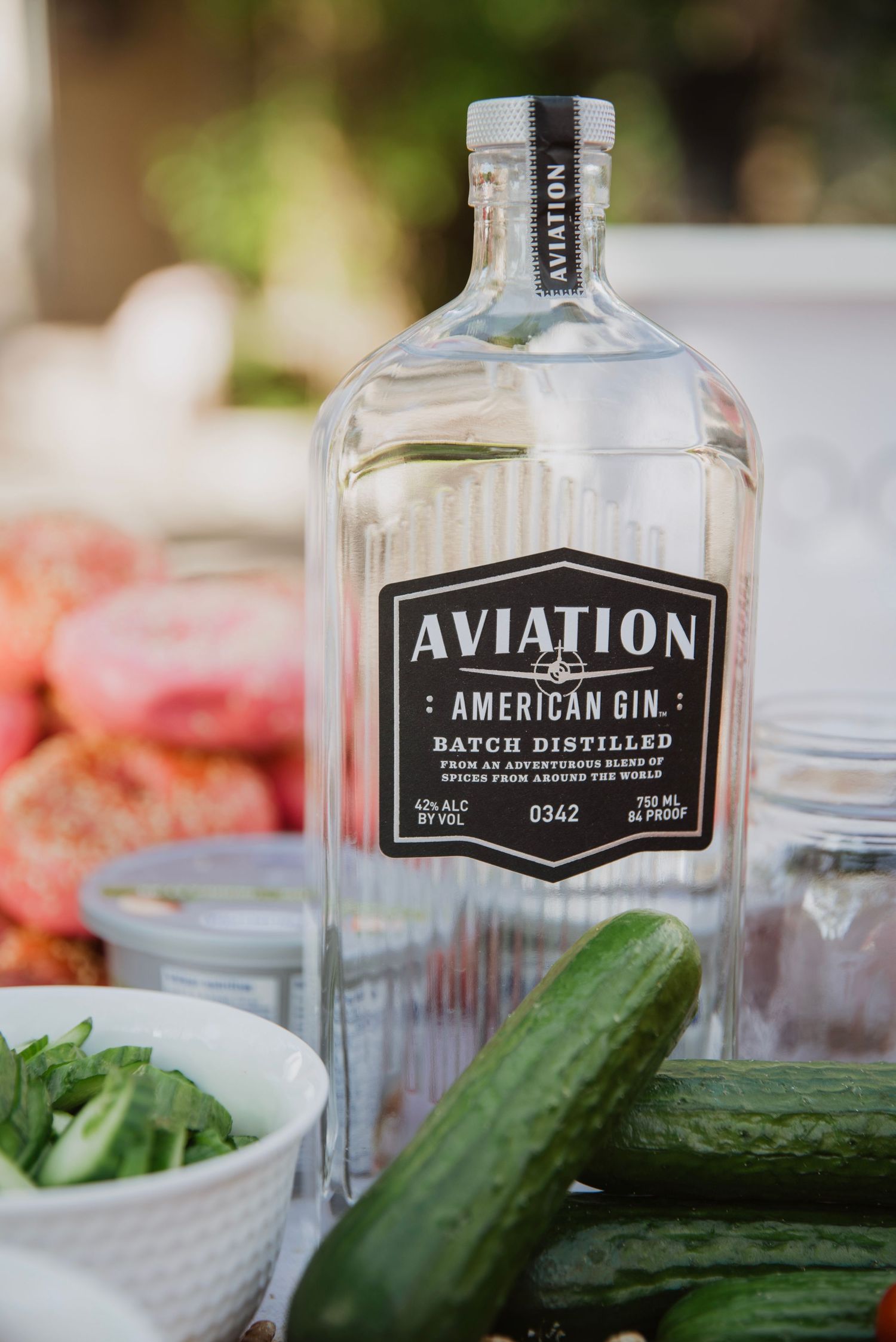 Aviation American Gin batch distilled bottle displayed with fresh cucumbers, tomatoes, and herbs on a table.