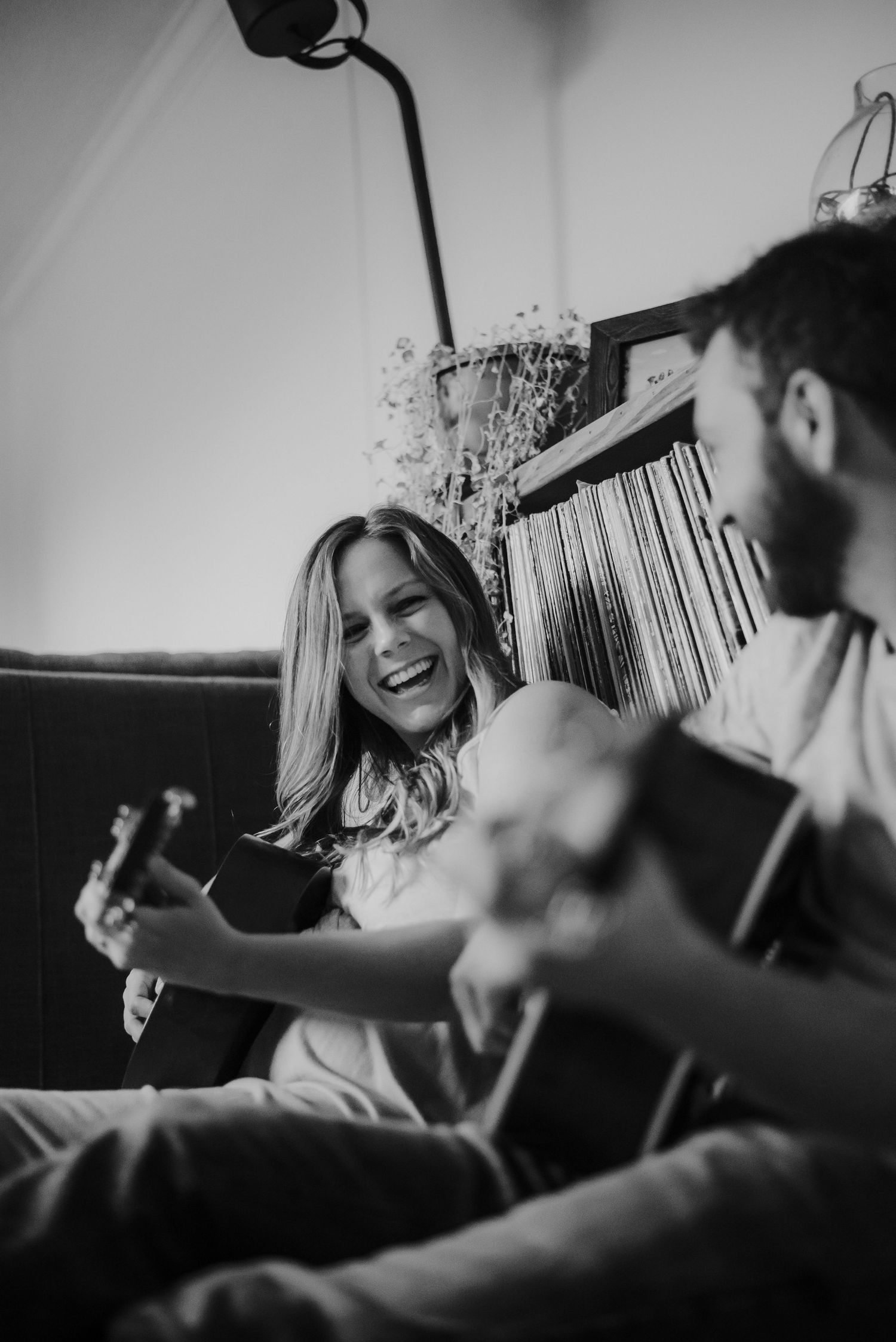 Black and white photo of a laughing woman playing ukulele with a man playing guitar on a couch.