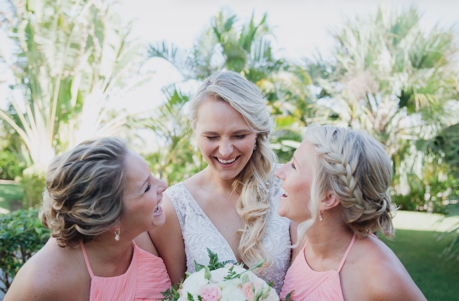 Bride in white lace gown laughing with two bridesmaids in pink dresses, holding bouquet outdoors.