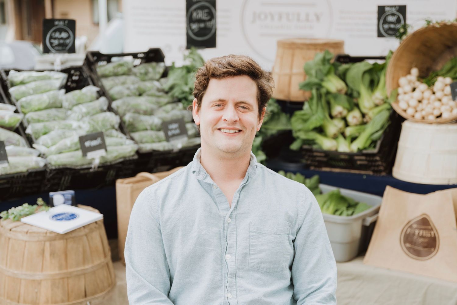 Smiling man at a farmers market stand with fresh vegetables and greens displayed behind him.