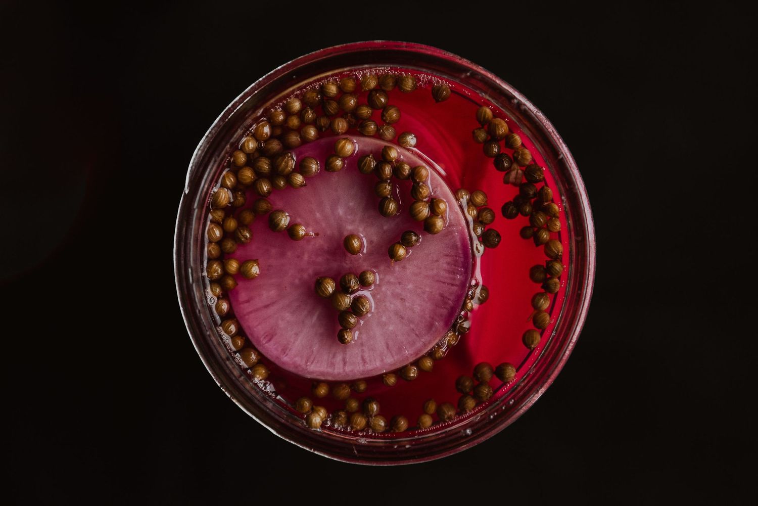 Top view of a vibrant red cocktail or mocktail with green peppercorns and a slice of beet on black background.