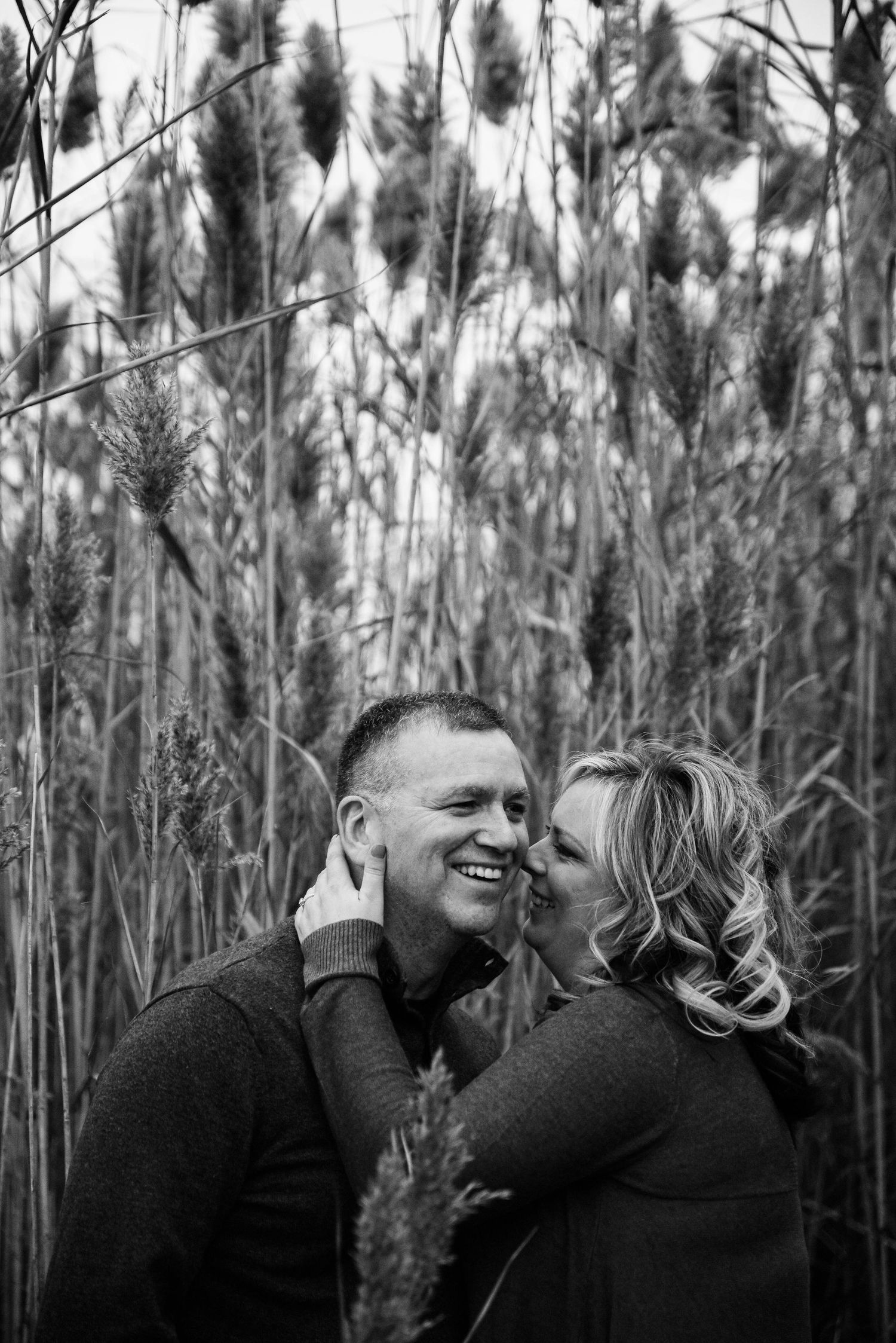 Black and white photo of a smiling couple embracing in tall grass, woman kissing mans cheek outdoors.