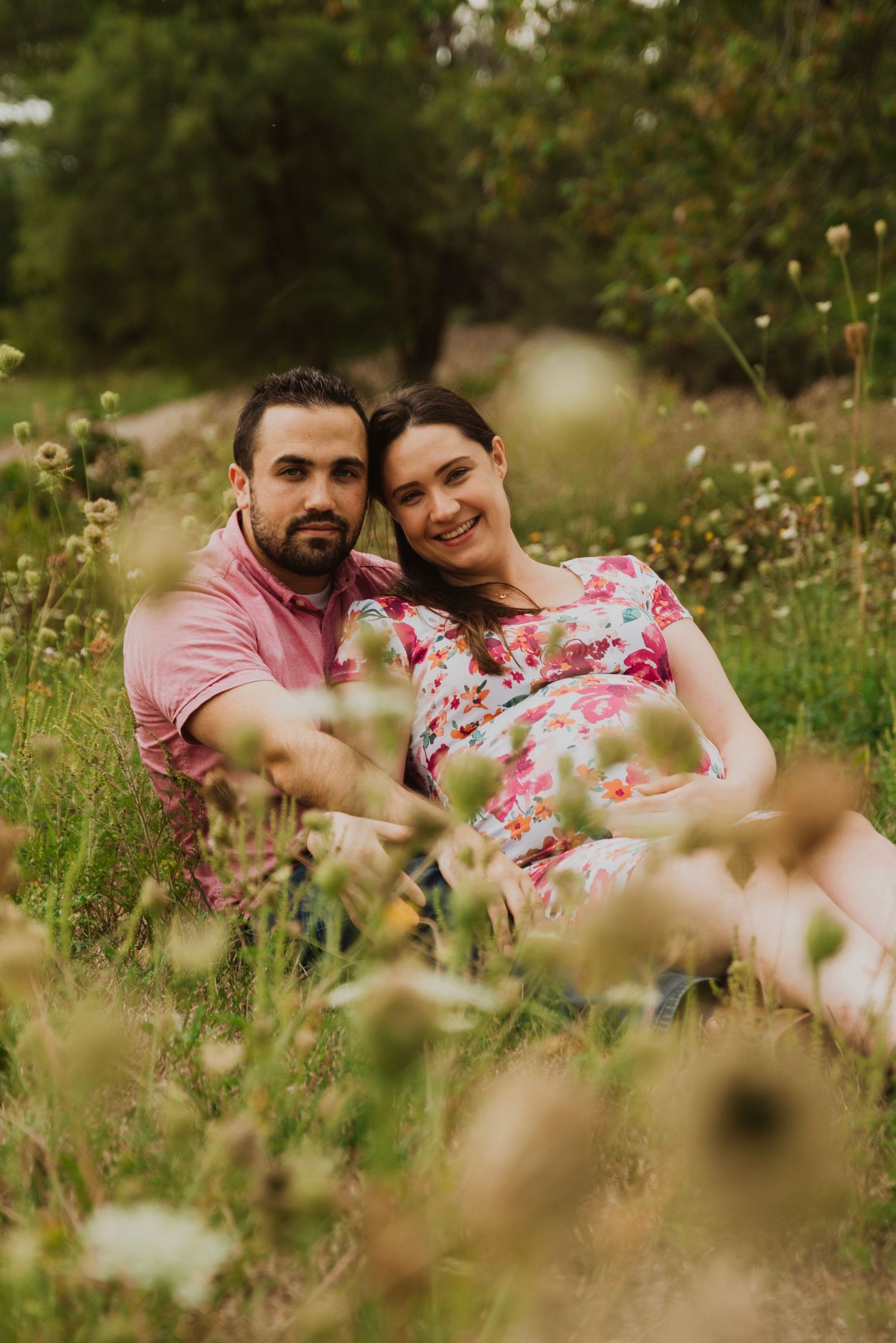 Couple sitting together in a wildflower meadow, woman in floral dress leaning on mans shoulder, smiling outdoors.