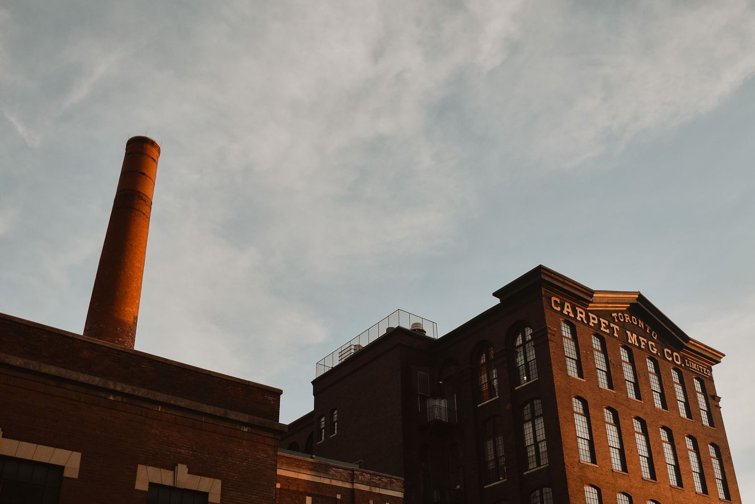 Historic brick factory buildings with tall red chimney stack against a blue sky, bathed in warm golden hour light.