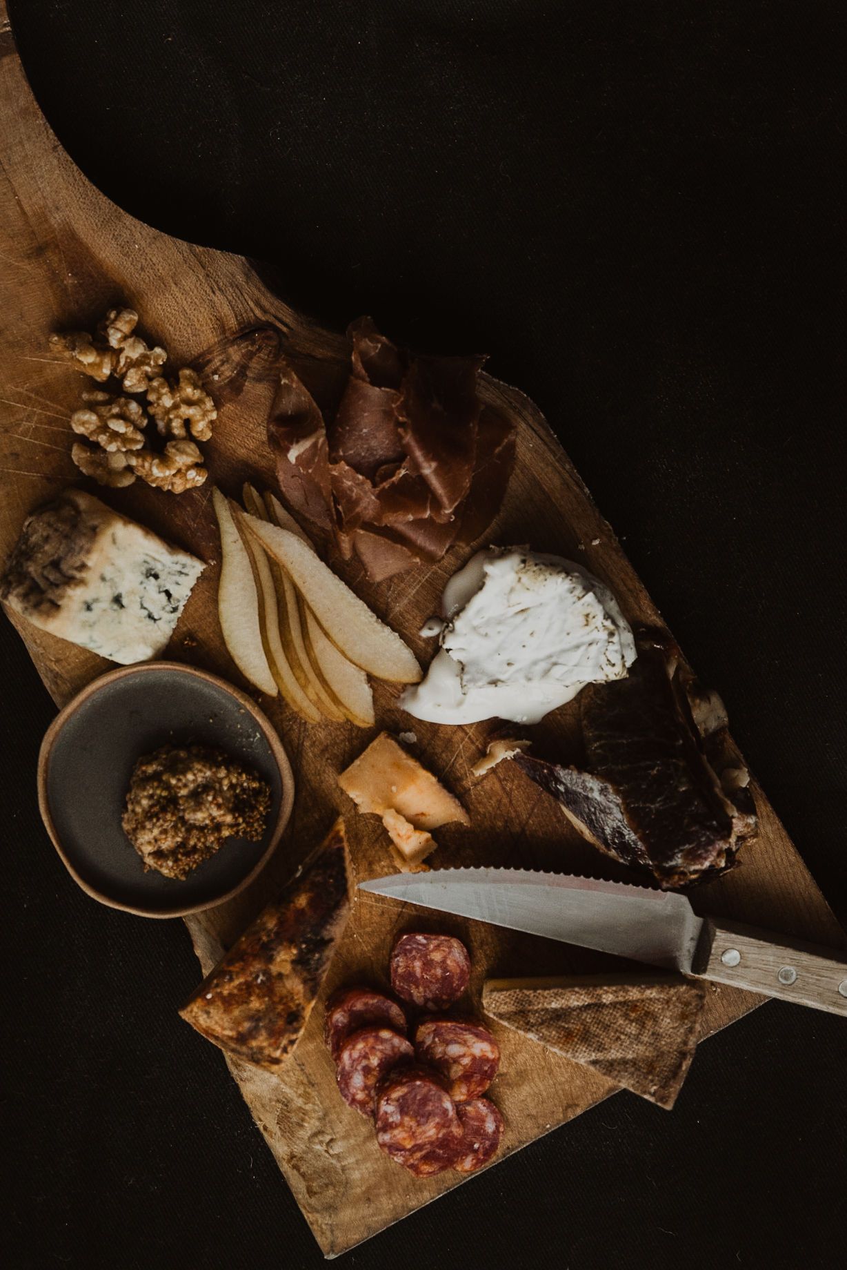Rustic charcuterie board with blue cheese, brie, cured meats, sliced pears, walnuts, salami, and a knife on dark background.