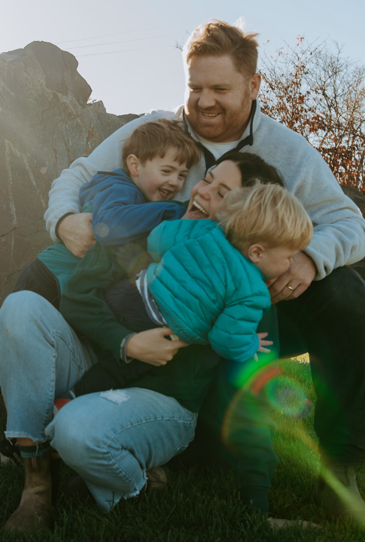 Happy family of four laughing and hugging outdoors on a sunny day, parents embracing two young boys.