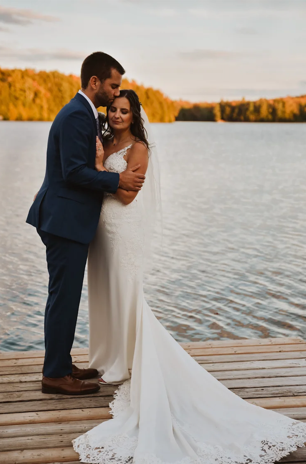 A couple in wedding attire embraces on a dock by a lake with autumn foliage at sunset.
