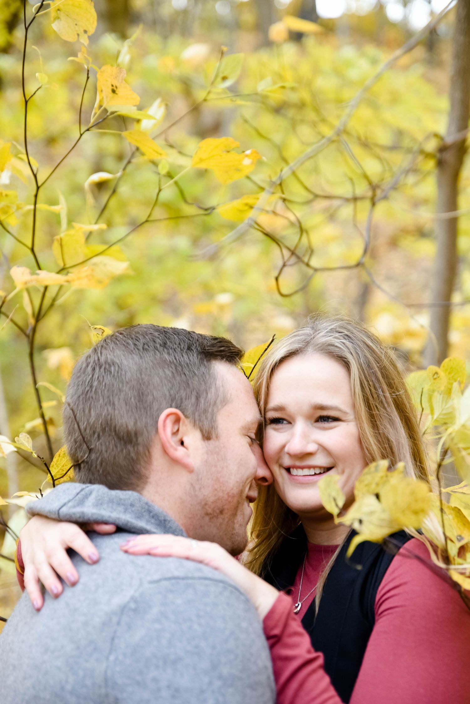 Couple sharing a romantic moment outdoors among yellow autumn leaves, woman smiling joyfully as man whispers in her ear.
