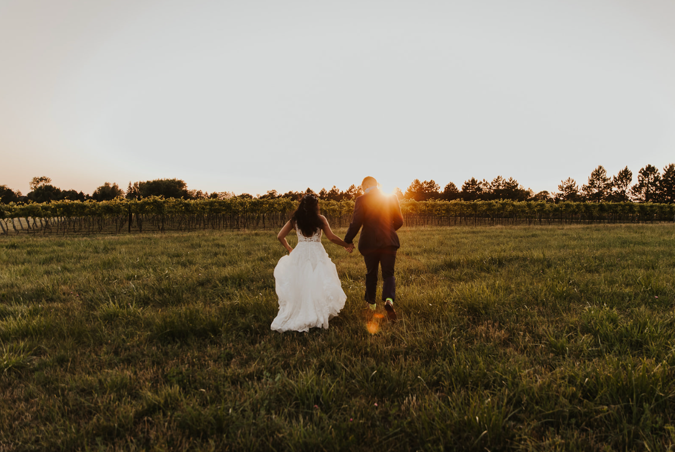 Bride and groom holding hands walking through vineyard at golden sunset hour in a romantic outdoor wedding photo.