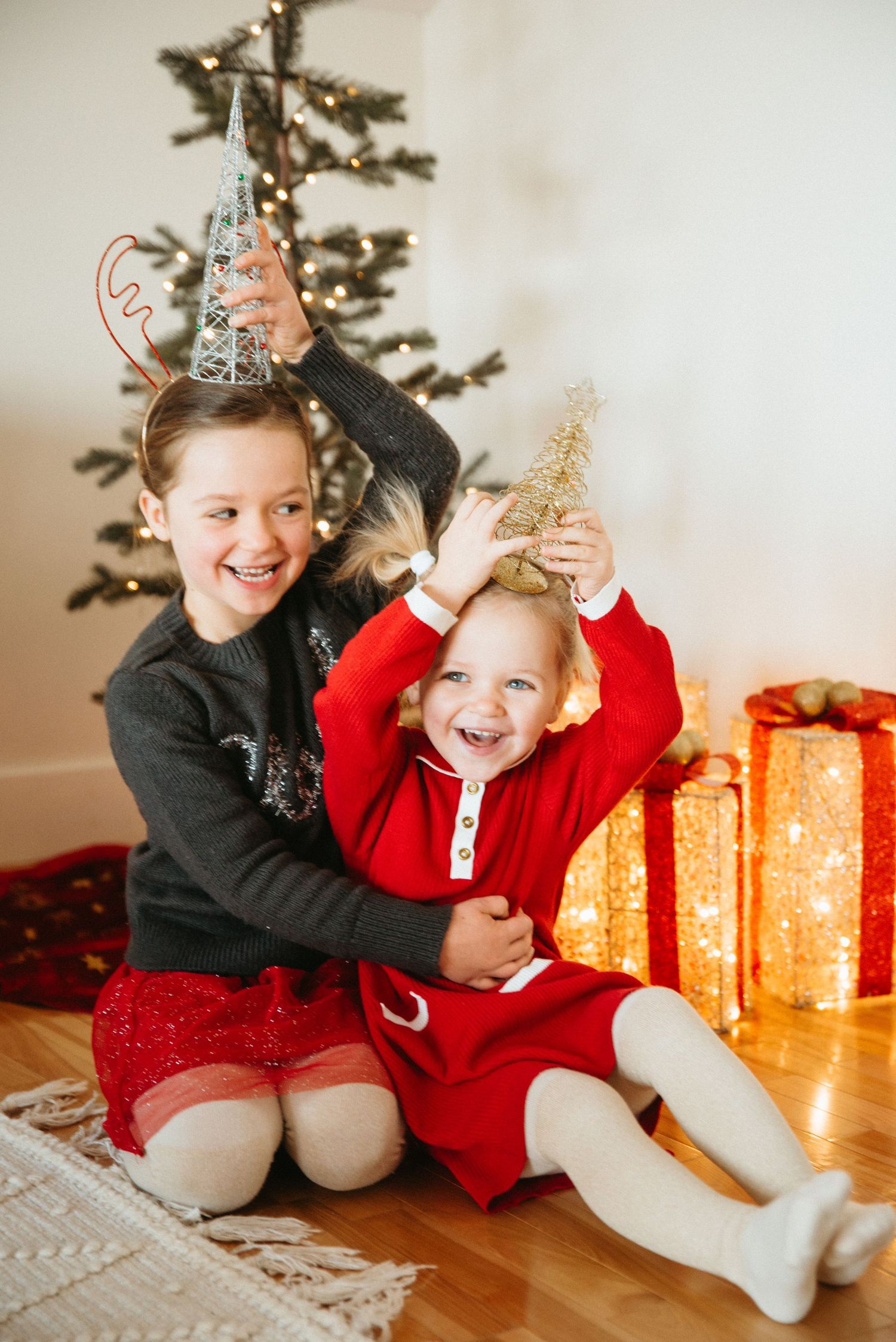 Two girls in Christmas outfits laughing and holding mini trees near a decorated Christmas tree with glowing gifts.