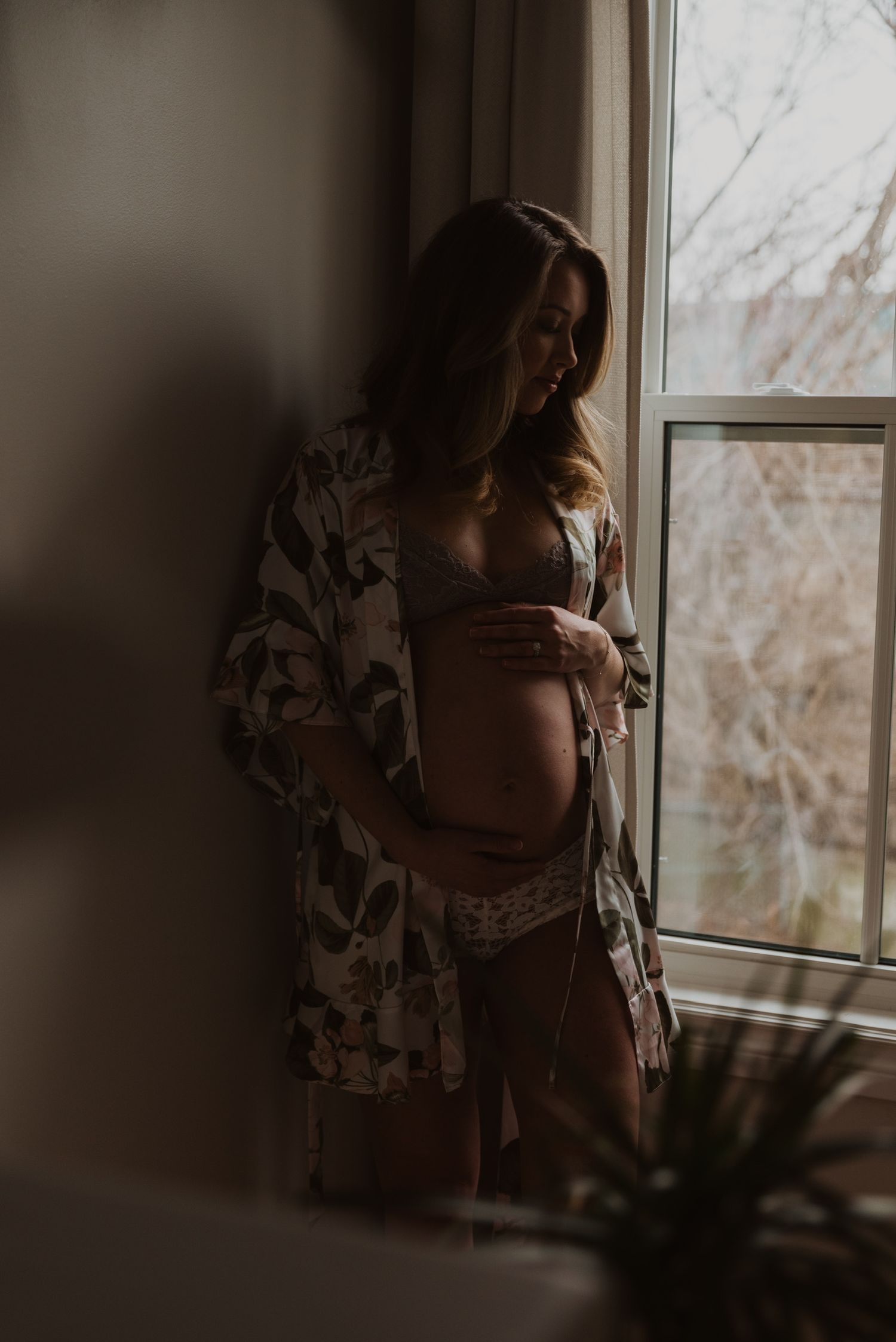 Pregnant woman in floral robe and lingerie cradling baby bump by a window in soft natural light.