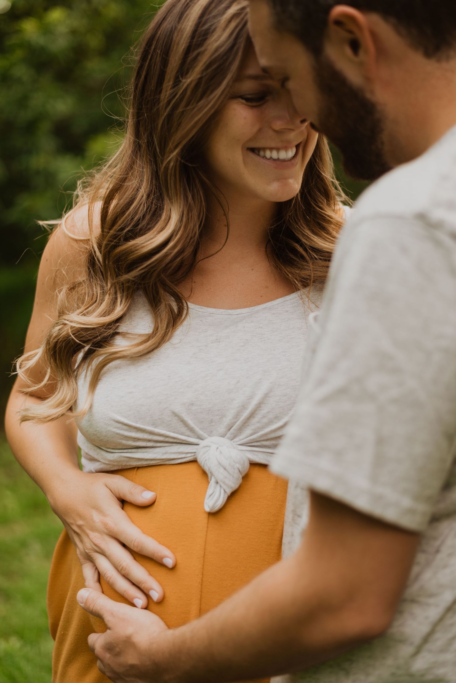 Smiling pregnant woman in white top and mustard skirt with partner touching her baby bump outdoors.