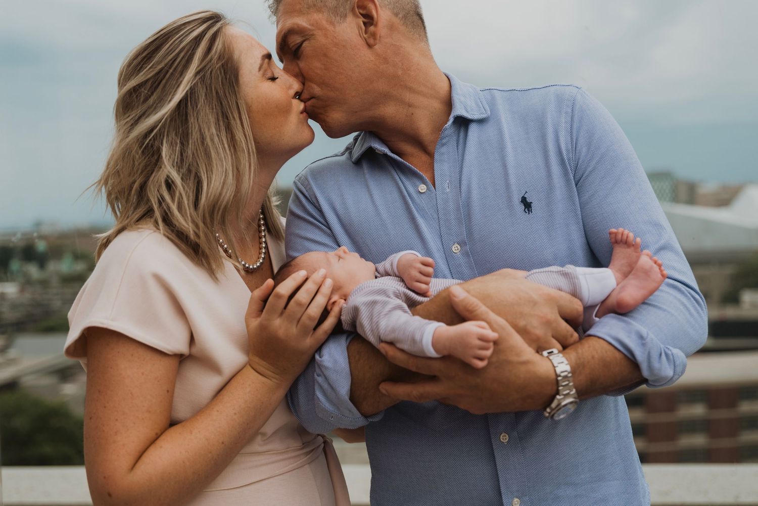 A couple shares a kiss while the man holds their newborn baby on an outdoor rooftop.