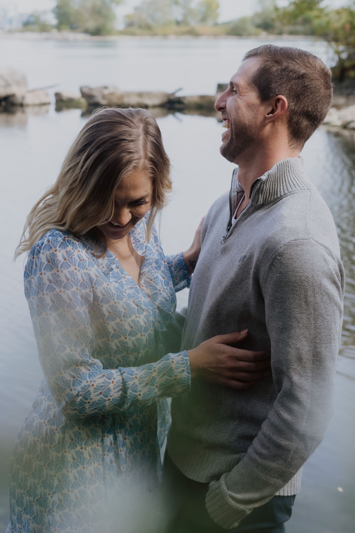 Couple sharing a tender moment by a lake, woman in blue lace dress, man in gray sweater, smiling together outdoors.