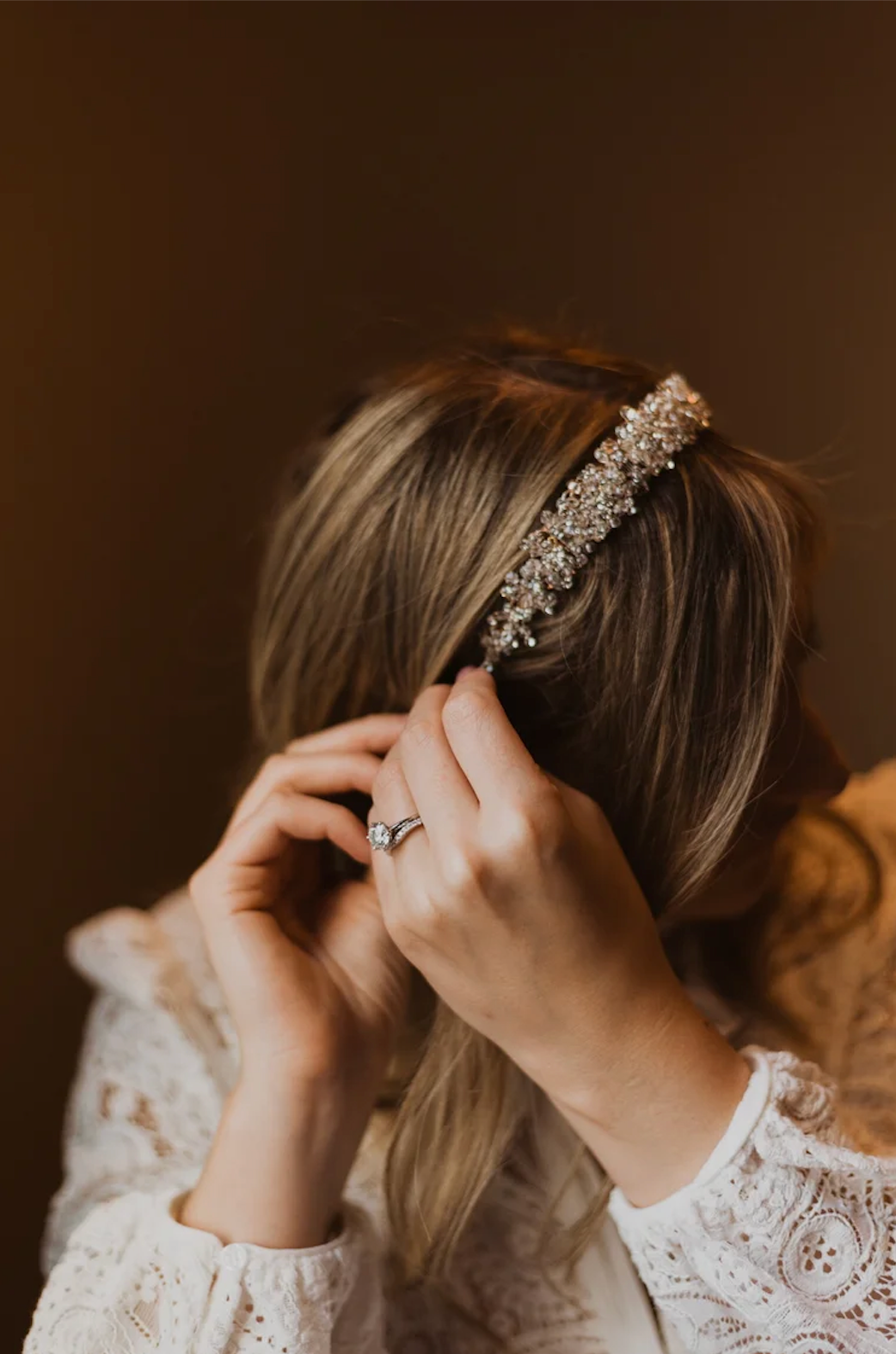 Bride placing a crystal embellished headband in her blonde hair, wearing a lace dress and diamond engagement ring.