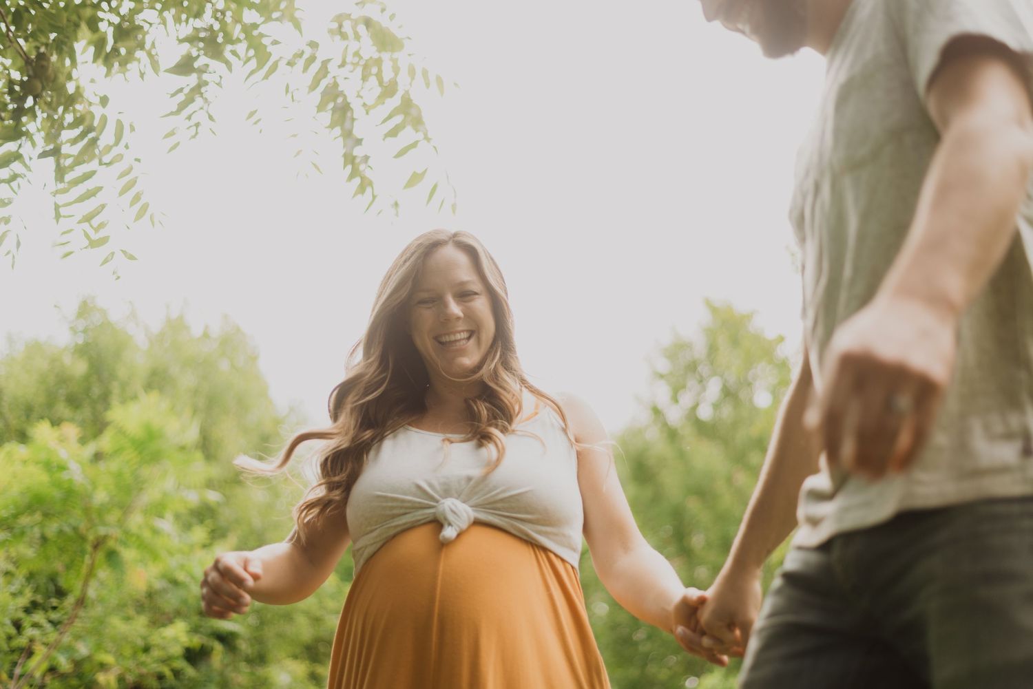 Smiling pregnant woman in white top and orange skirt holds hands with partner outdoors among lush green trees.