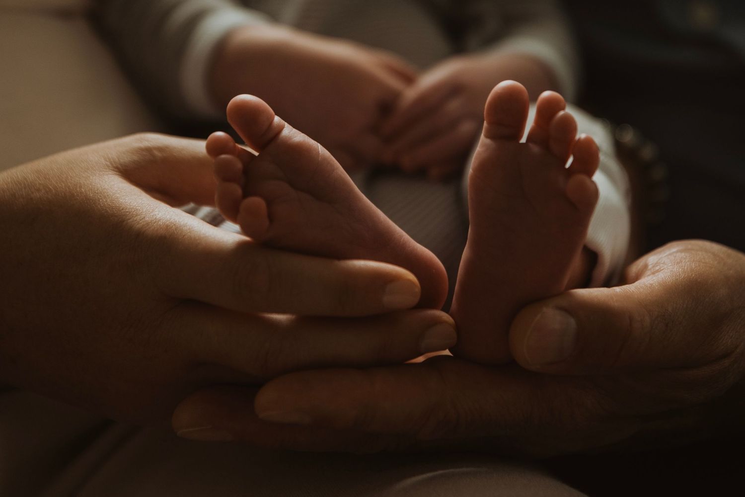 Adult hands gently cradling tiny newborn baby feet in a warm, intimate close-up photo.