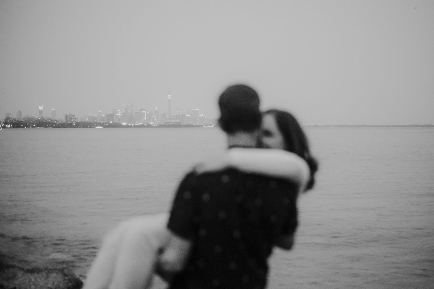 Black and white photo of a couple embracing by the waterfront with a city skyline in the misty background.