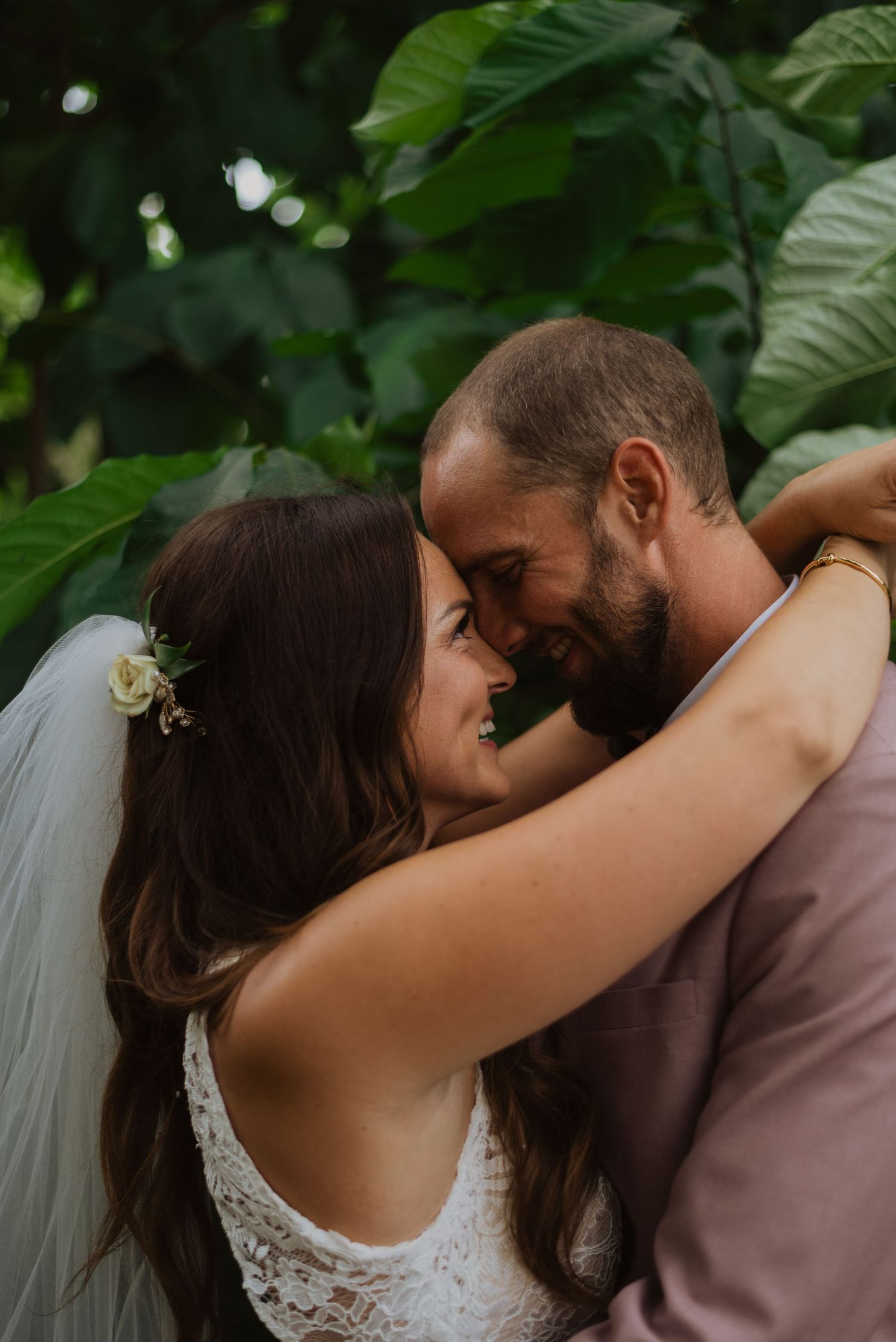 A bride and groom share an intimate moment, touching foreheads and smiling, surrounded by lush green foliage.