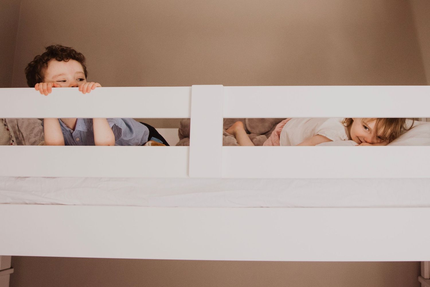 Two young children peeking over the rails of a white bunk bed, smiling and playing together.