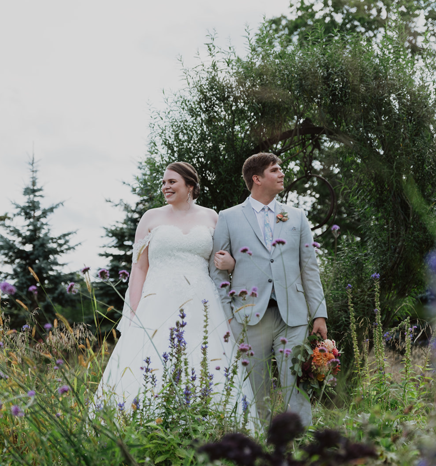 A bride and groom walk arm in arm through a wildflower garden, dressed in a white gown and light gray suit.