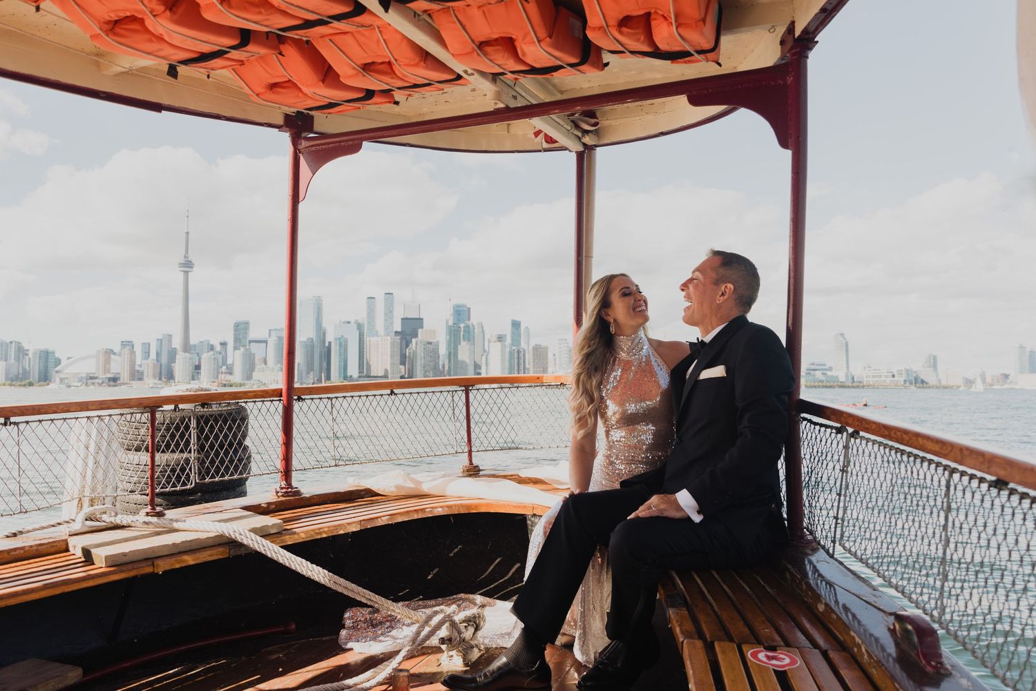 Couple in wedding attire sitting on a vintage boat with Toronto skyline and CN Tower in the background.