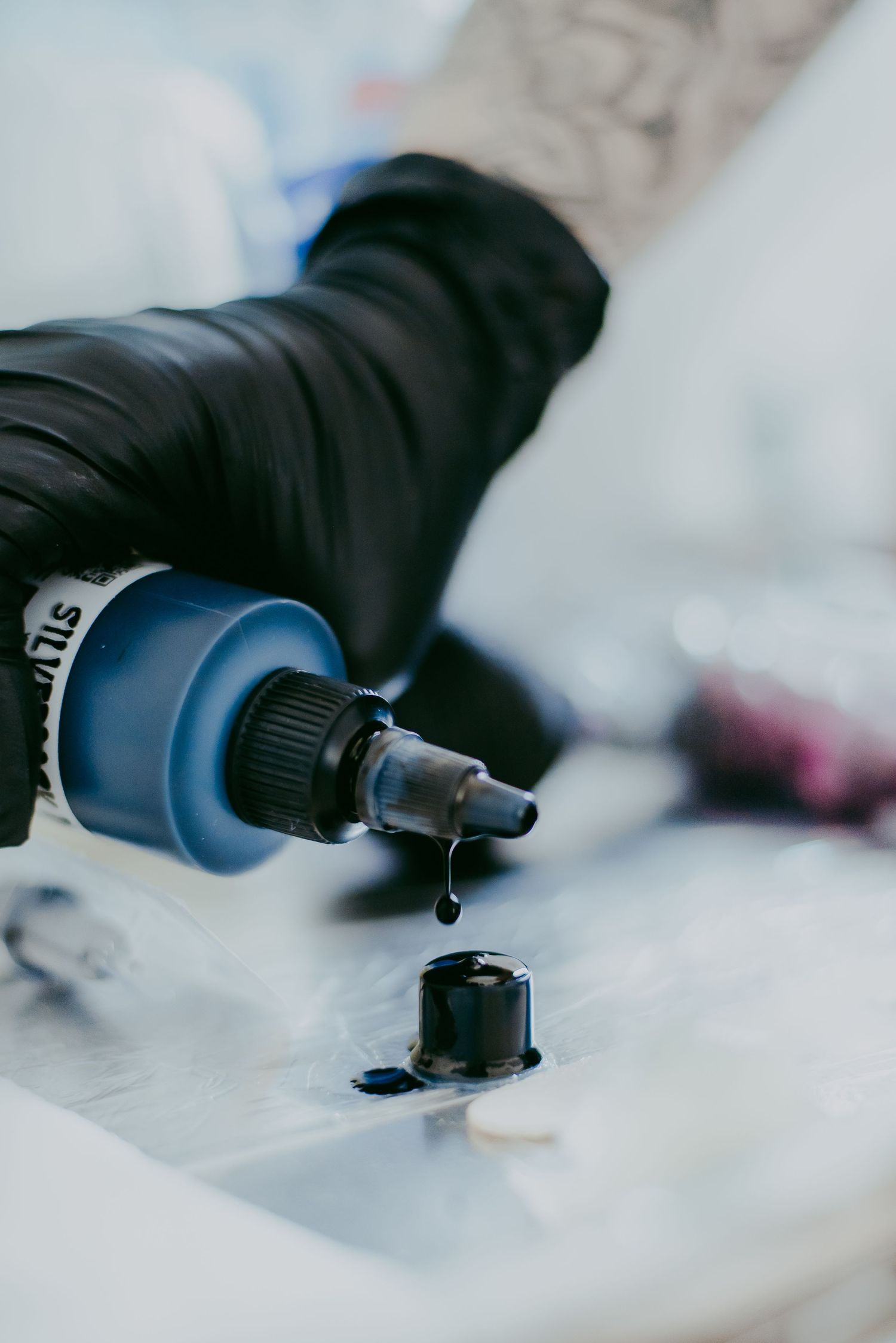 Tattoo artist in black gloves pouring dark ink from a blue bottle into a small cap on a white surface.