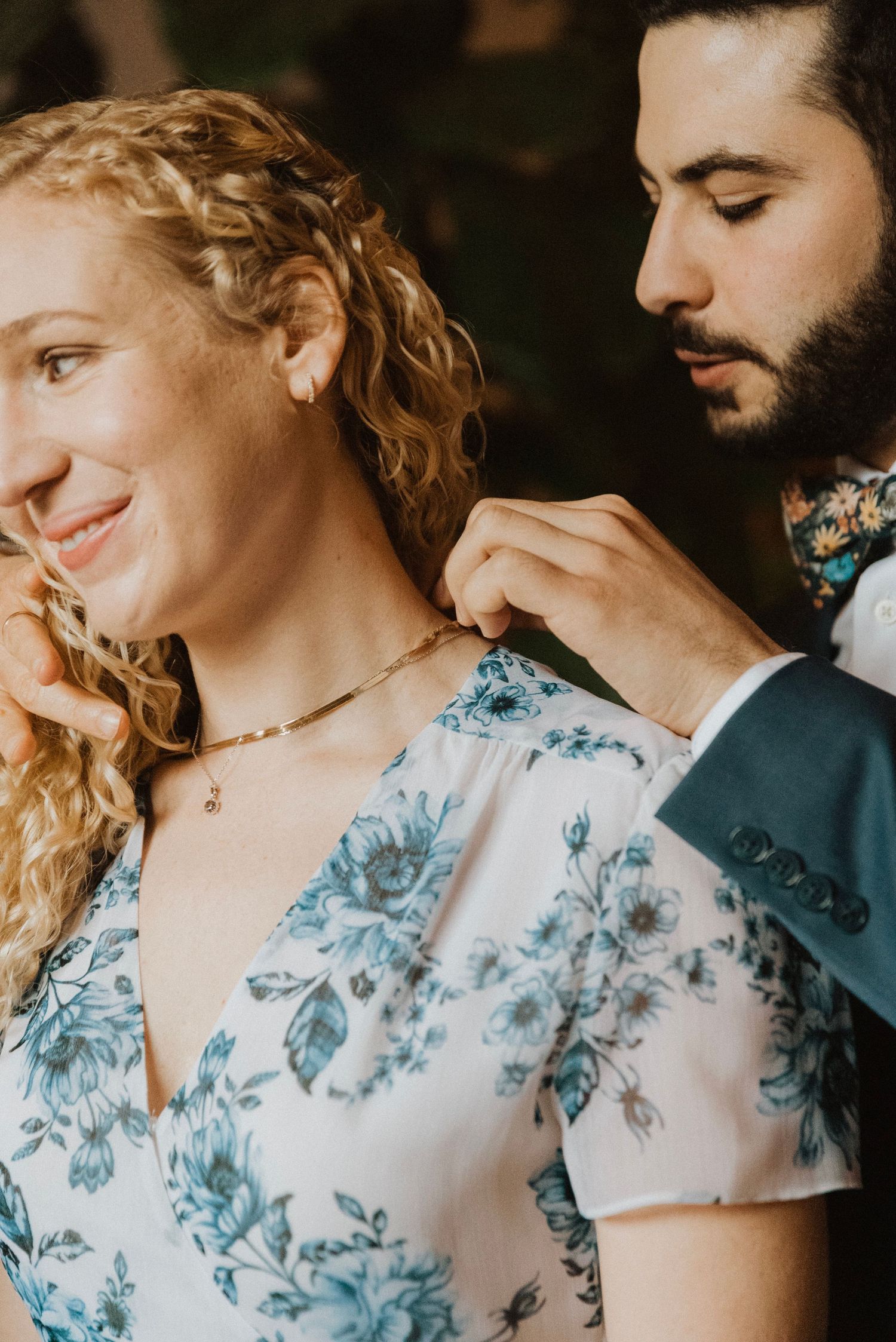 A man with a beard fastens a delicate gold necklace on a smiling woman in a floral blue dress.