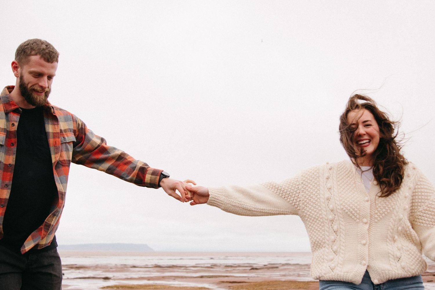 Happy couple holding hands on a beach, man in plaid shirt and woman in cream knit sweater, laughing outdoors.